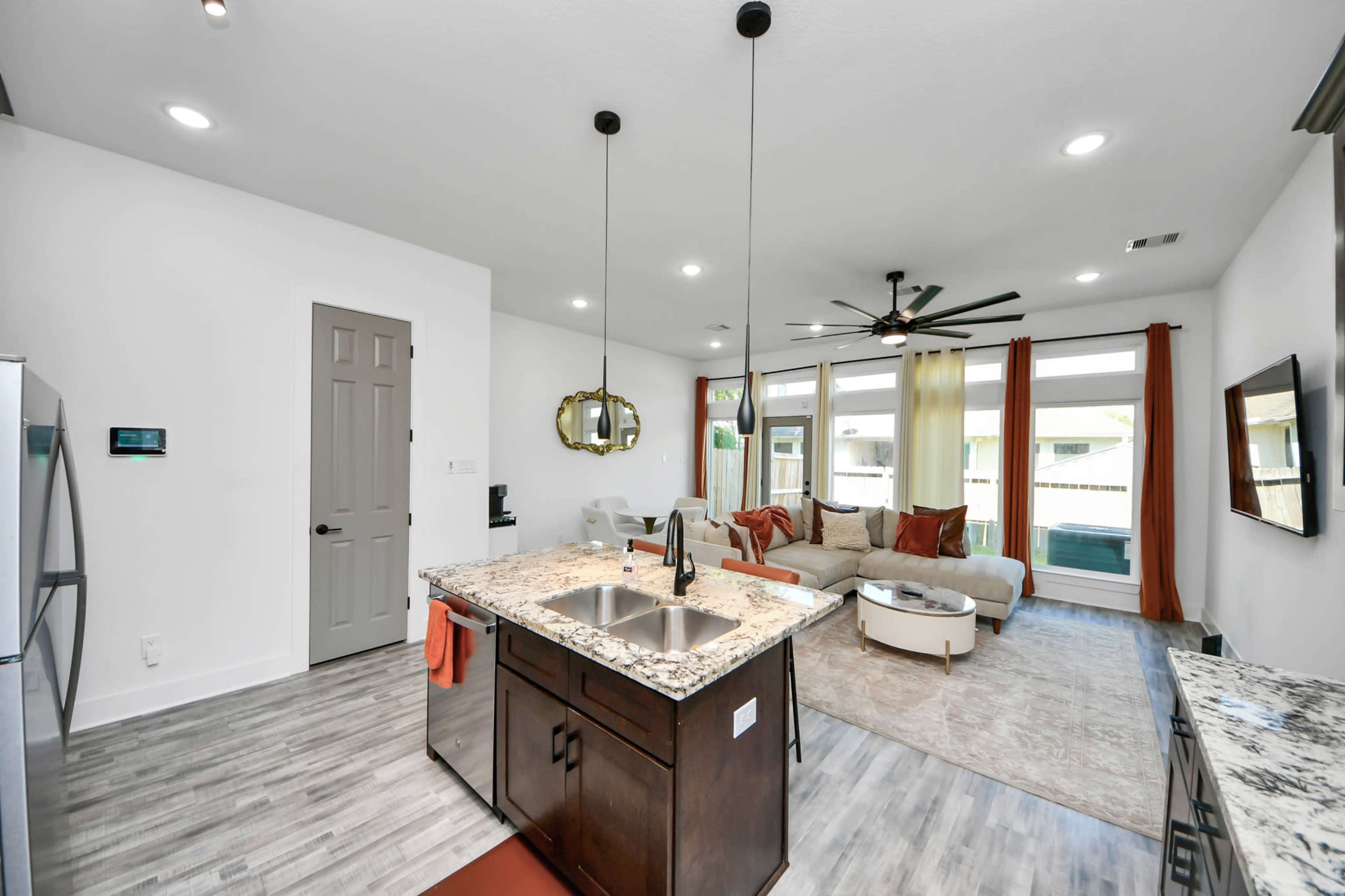 The image shows a modern open-concept kitchen and living area with granite countertops, pendant lights, and large windows allowing natural light to fill the space.