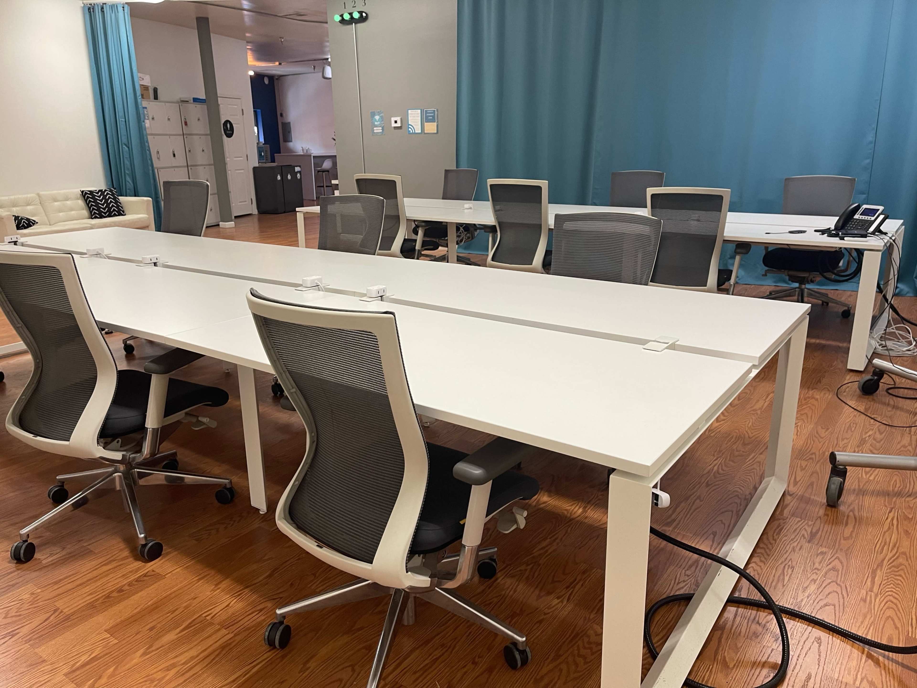 A modern office meeting room features a long white table surrounded by ergonomic chairs, with blue curtains and a phone on a side table.