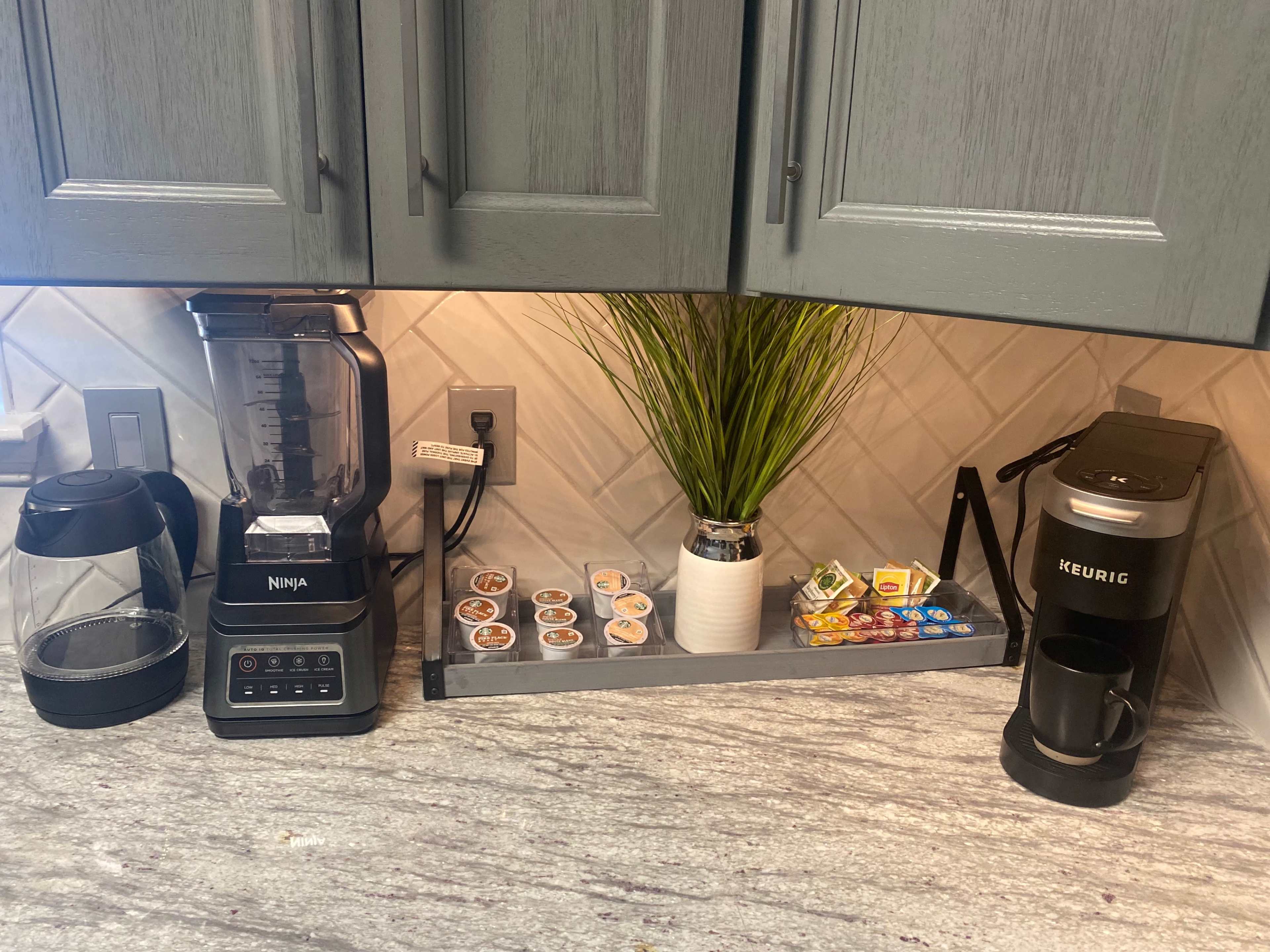 The image shows a kitchen countertop with a Ninja blender, a Keurig coffee maker, a tray of coffee pods, and a decorative plant in a white vase.