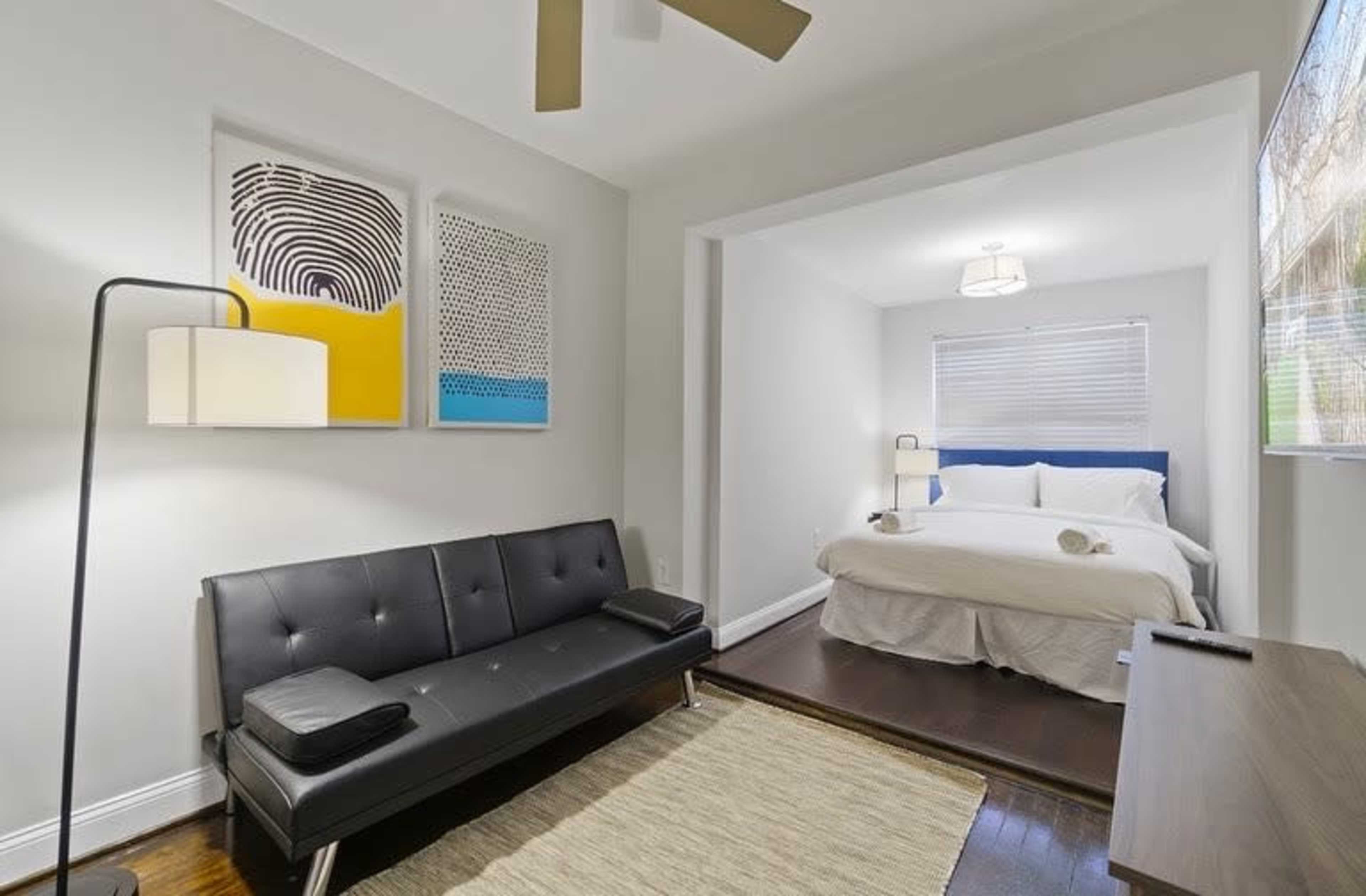 The image shows a modern bedroom featuring a bed, a black sofa, artwork on the walls, and a wooden dresser.