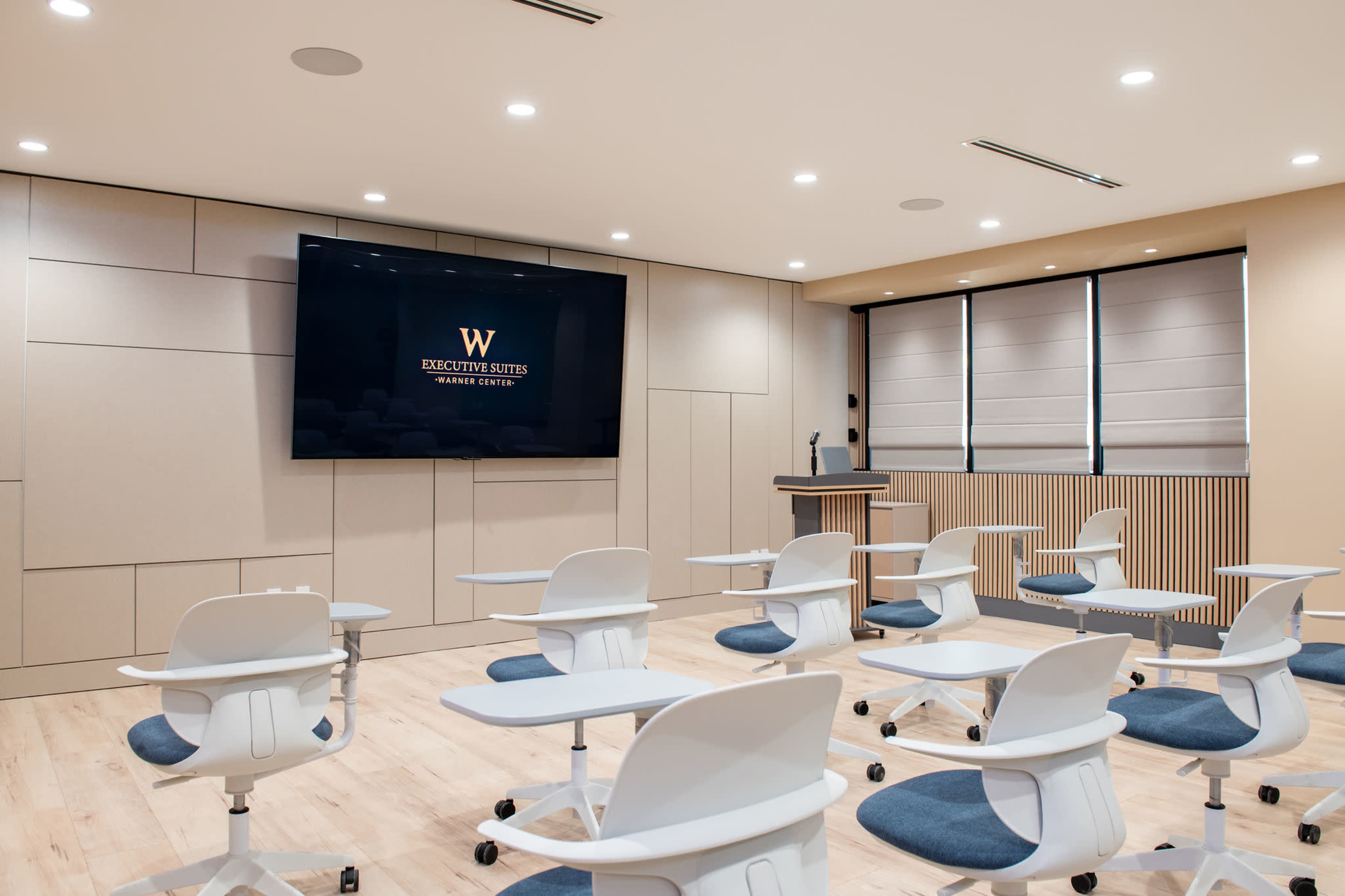The image shows a modern classroom featuring rows of adjustable white chairs with attached desks, a podium, and a large screen displaying a logo on the wall.