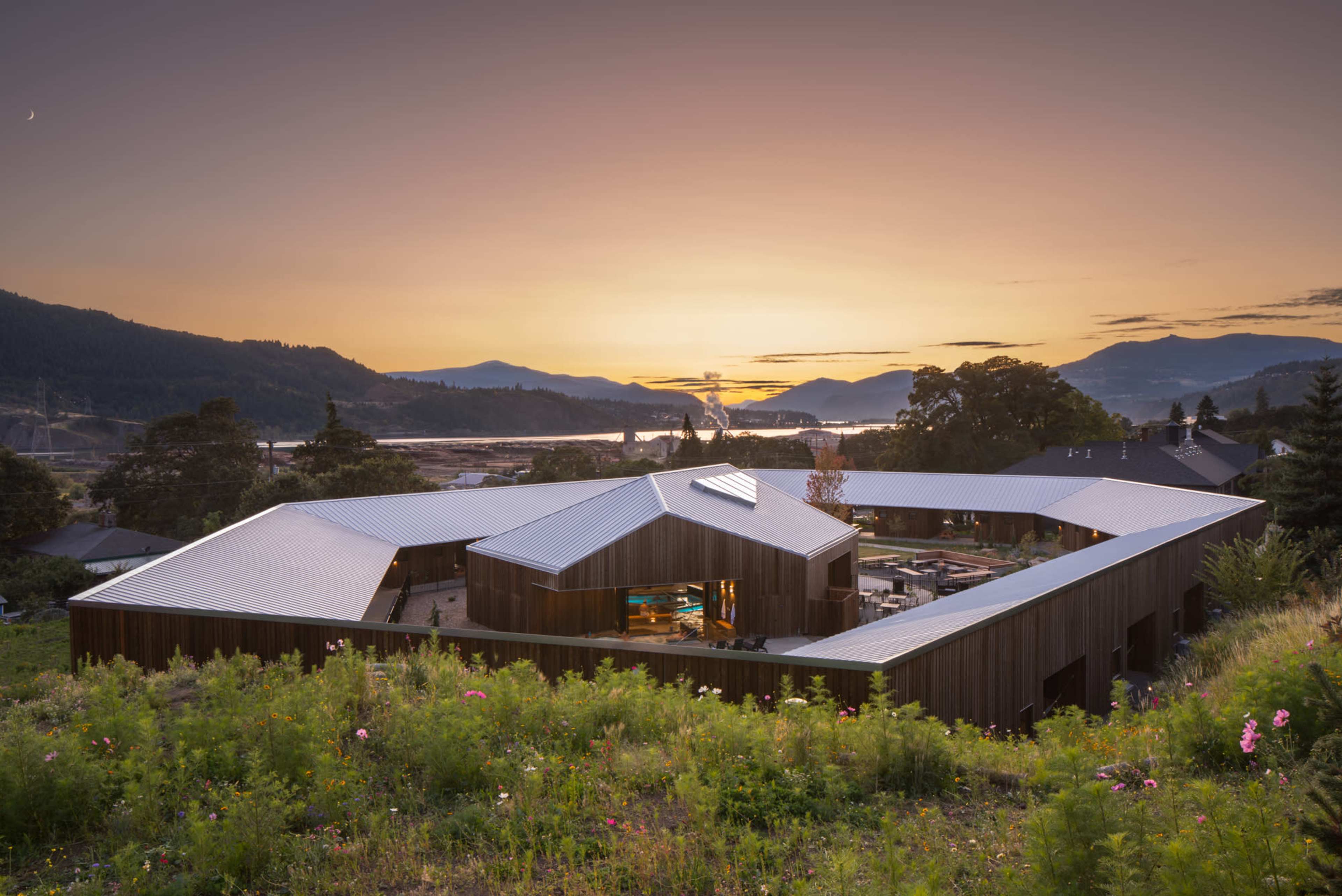 A modern wooden house with a unique asymmetrical design sits on a hillside overlooking a river and mountains at sunset.