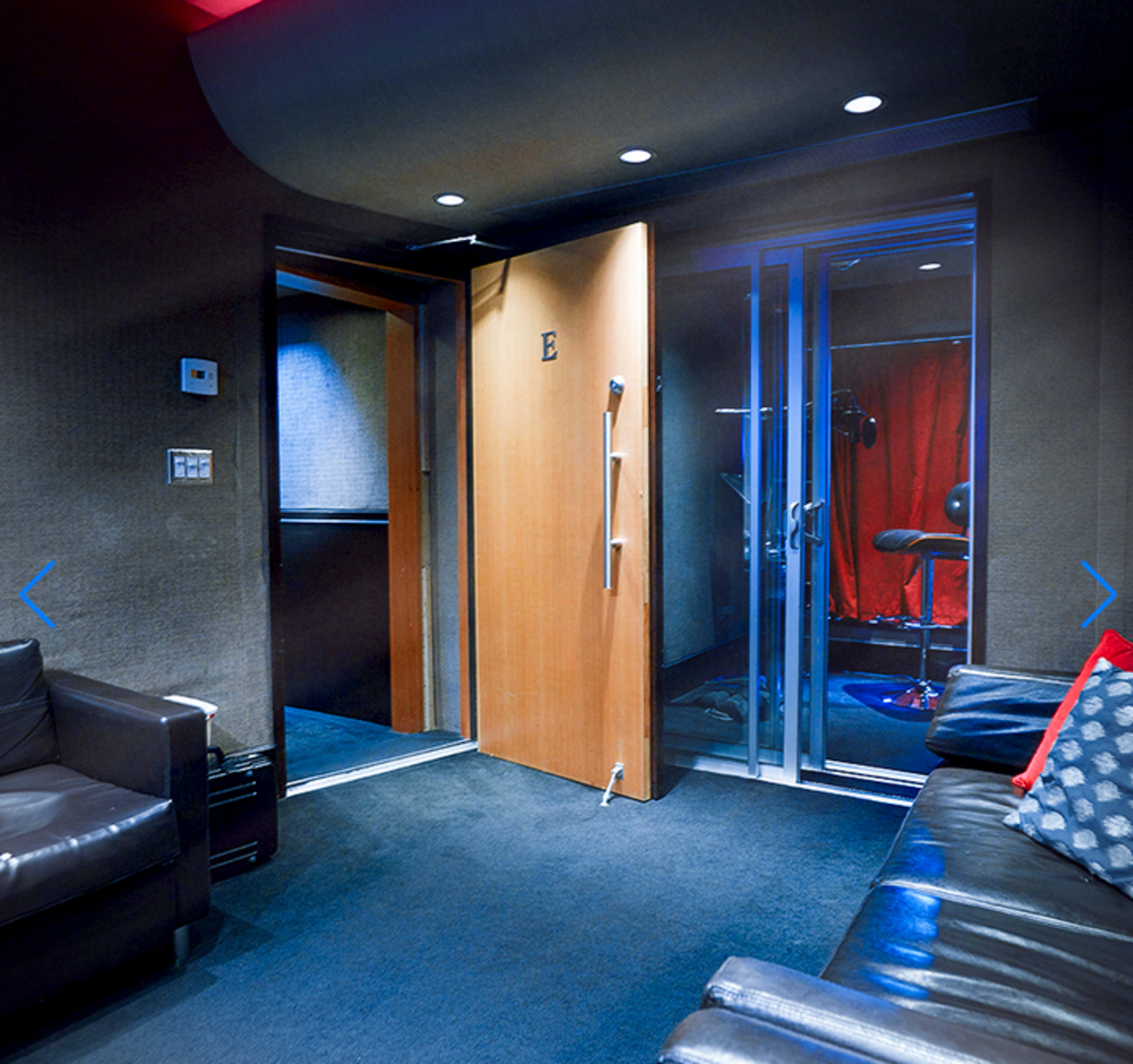 A modern room with dark walls, a partially open wooden door leading to a hallway, and glass doors that likely open to a soundproof booth.