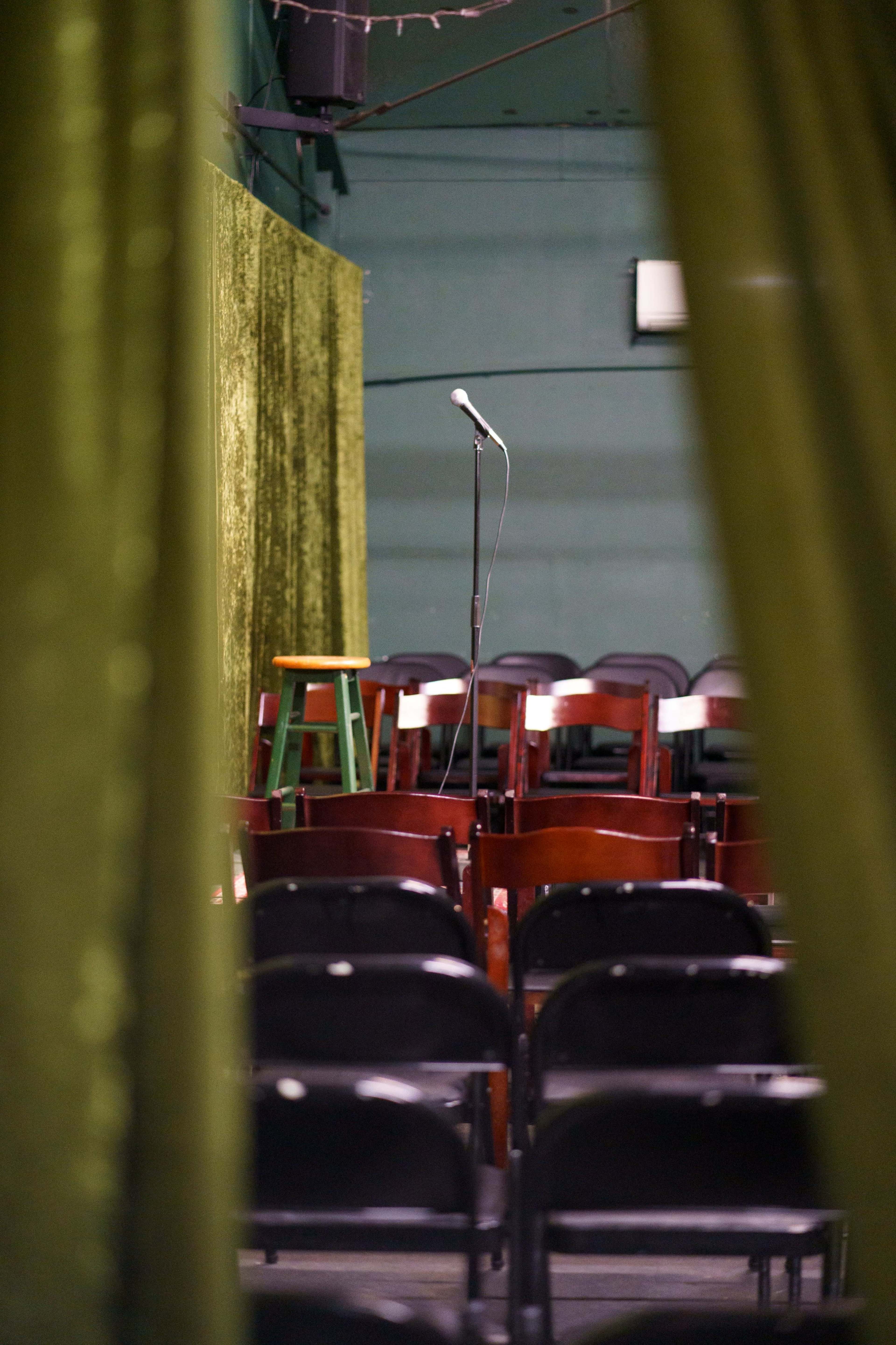 A microphone stands on a stool at the front of a small stage surrounded by rows of empty chairs.