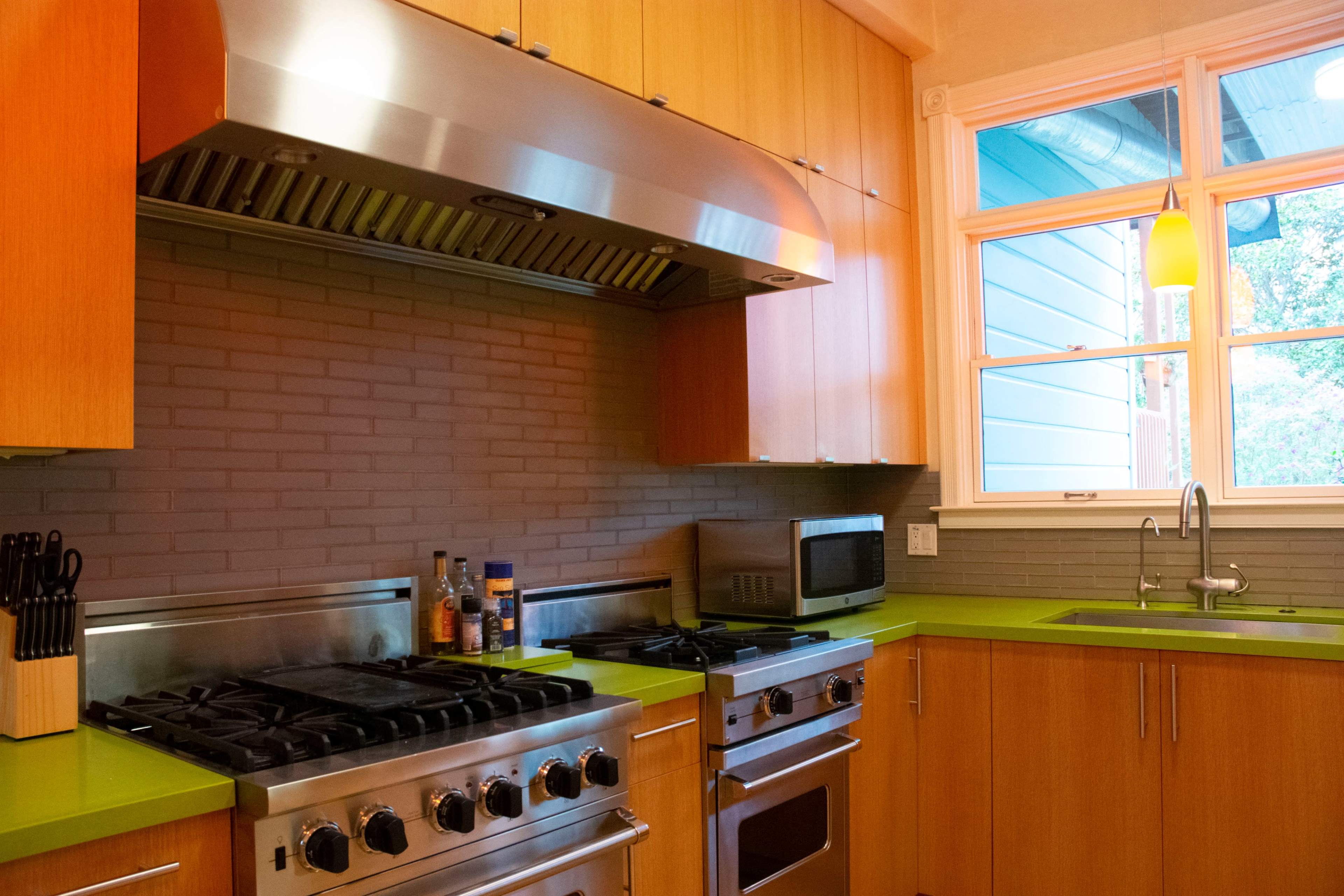A modern kitchen features wooden cabinetry, a stainless steel range and hood, and vibrant green countertops beside a window.