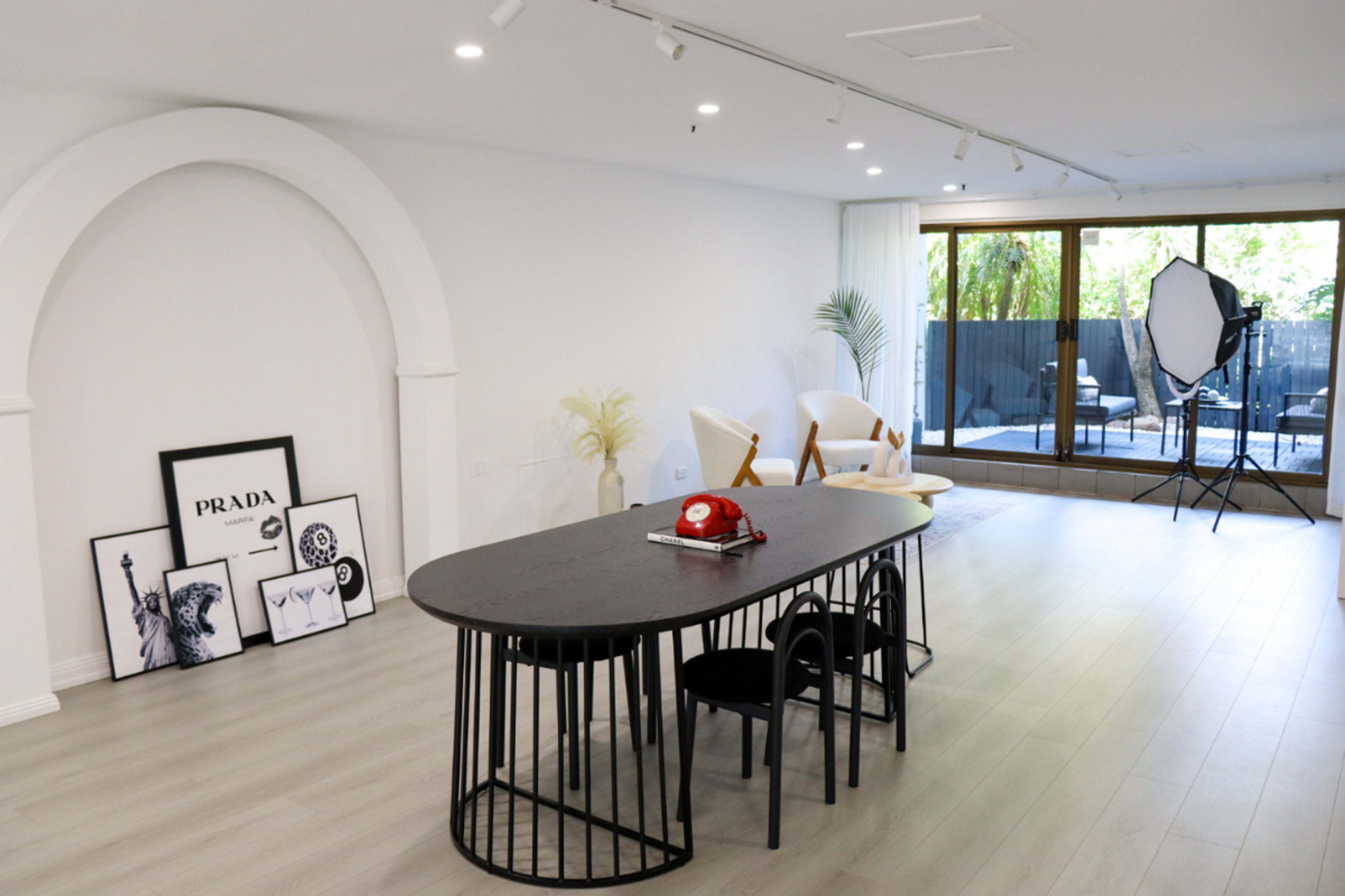 The image shows a modern, spacious room with a black dining table and chairs, framed artwork on the floor, and large windows leading to a patio.