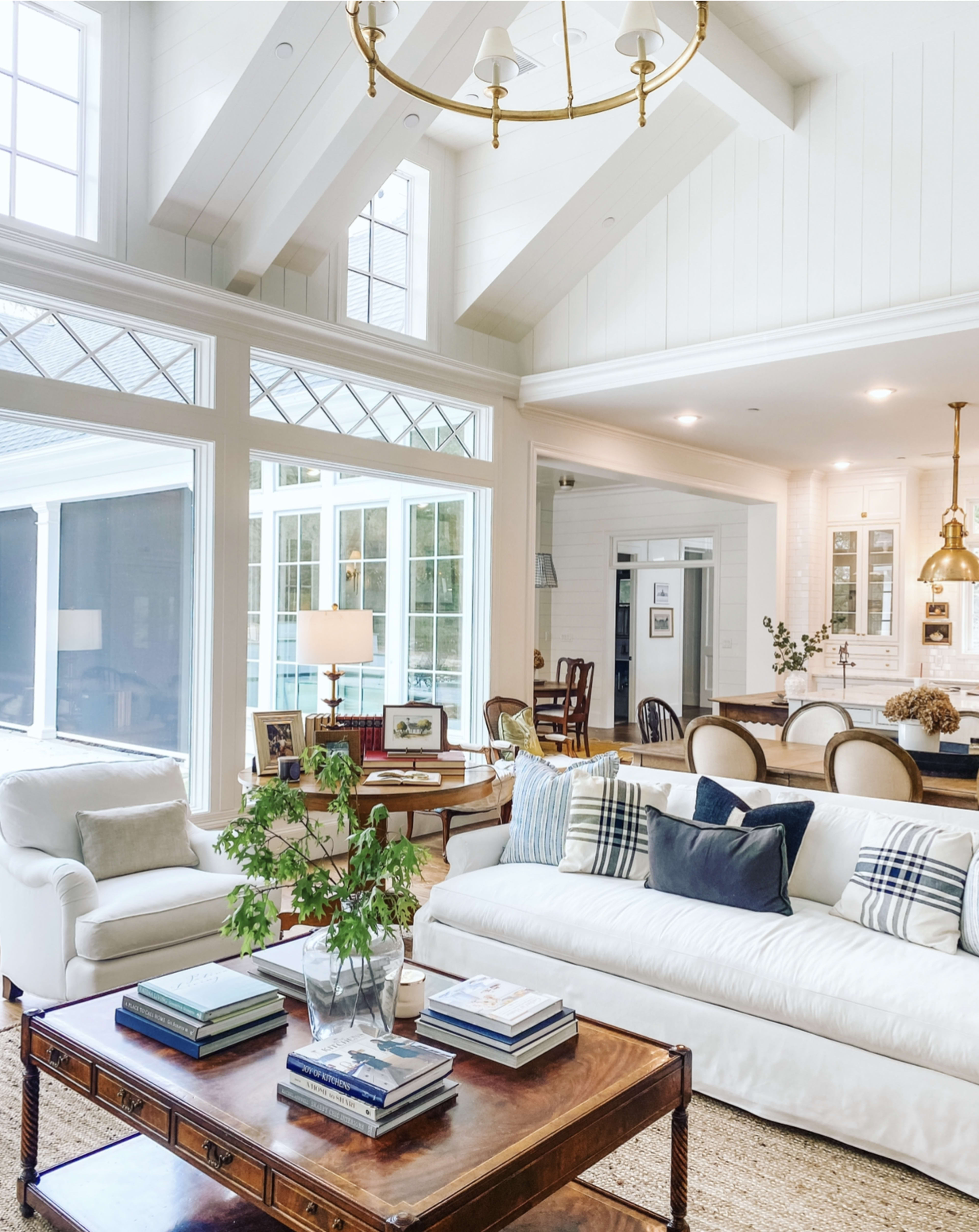 The living room features a white sofa, a wooden coffee table stacked with books, and large windows allowing sunlight to illuminate the space.