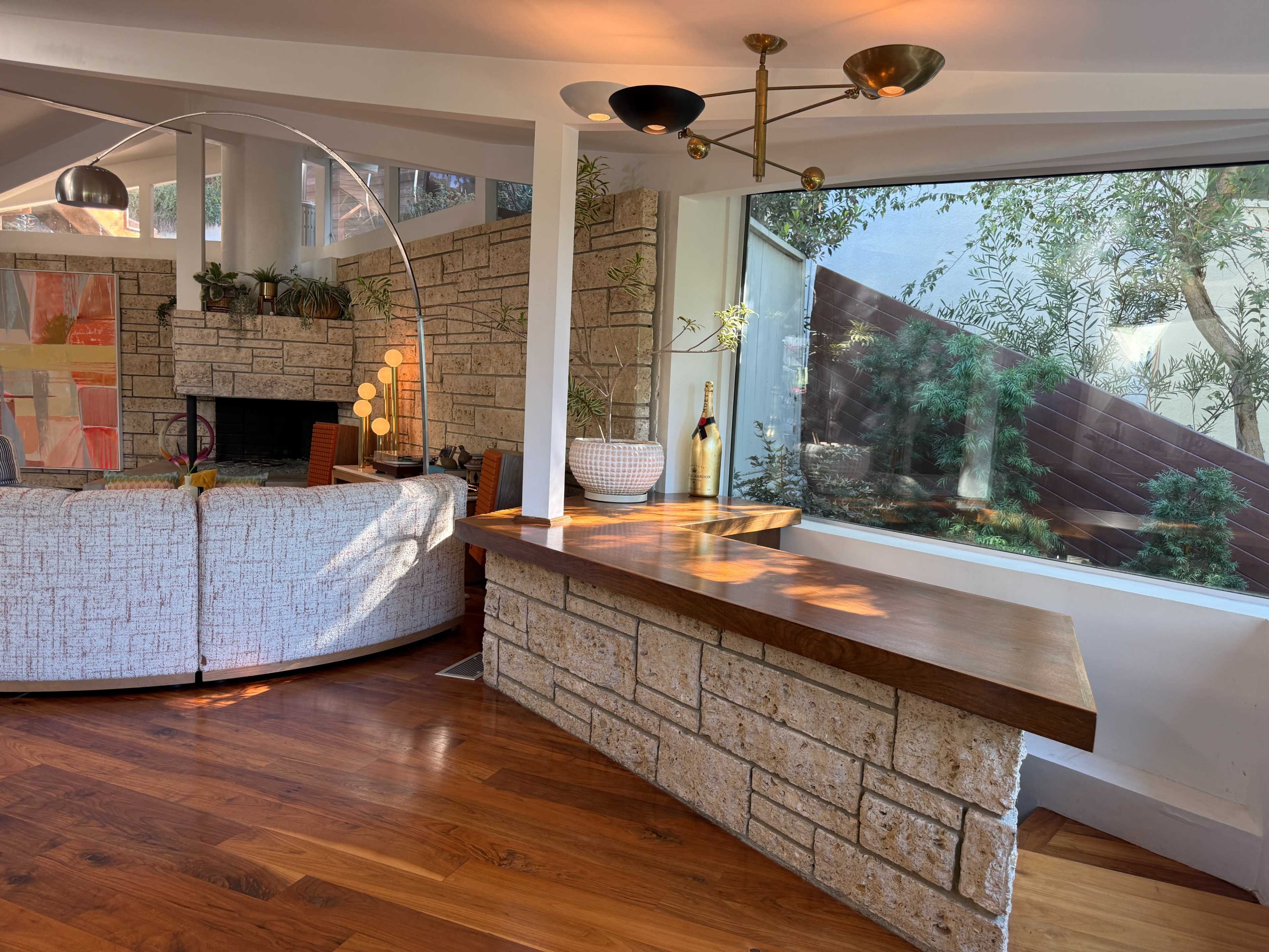 The image shows a modern interior space featuring a stone wall, a wooden countertop, a curved sofa, and large windows overlooking greenery.