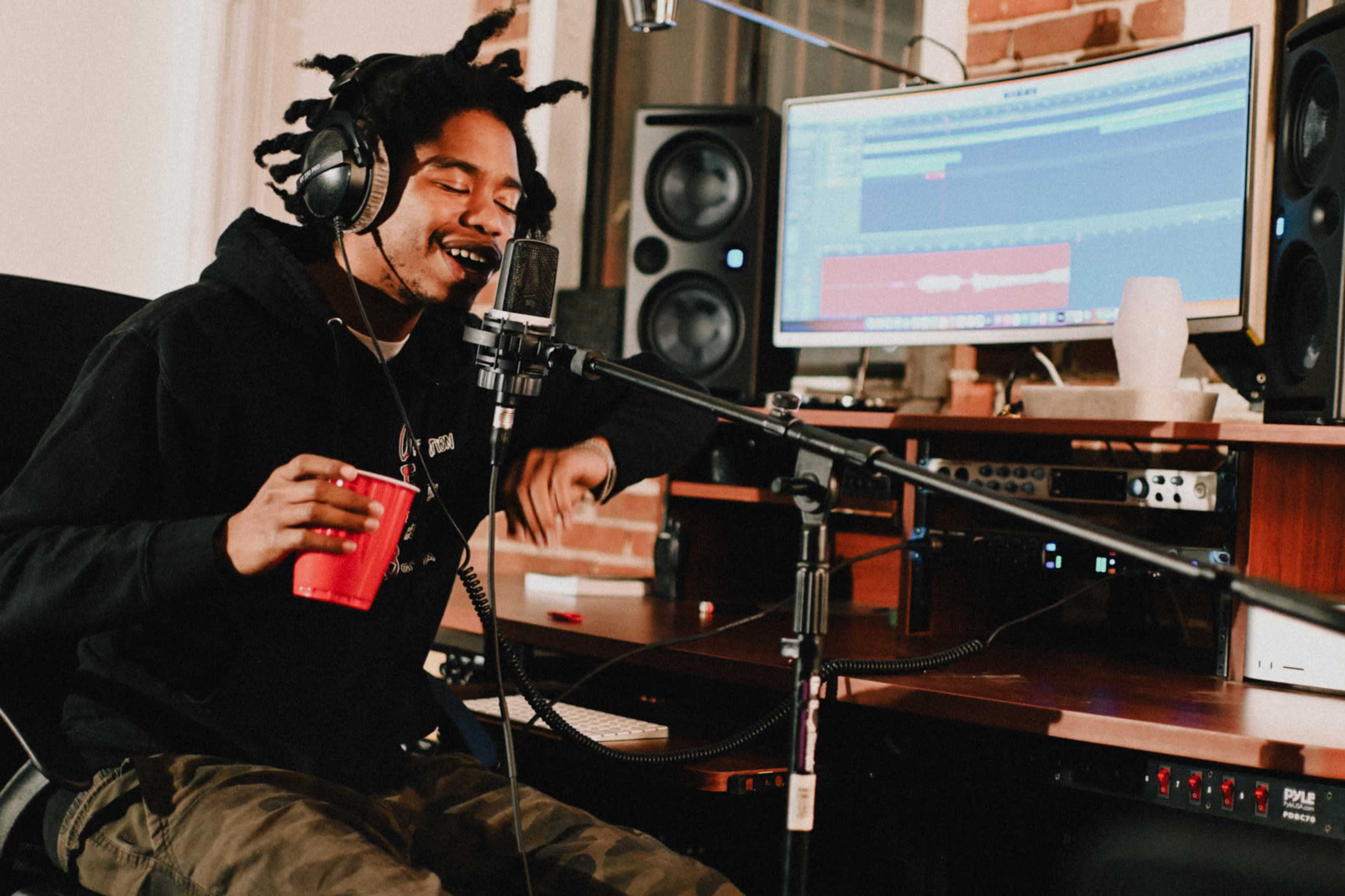 A musician wearing headphones records vocals while holding a red cup in a studio equipped with a computer and audio equipment.