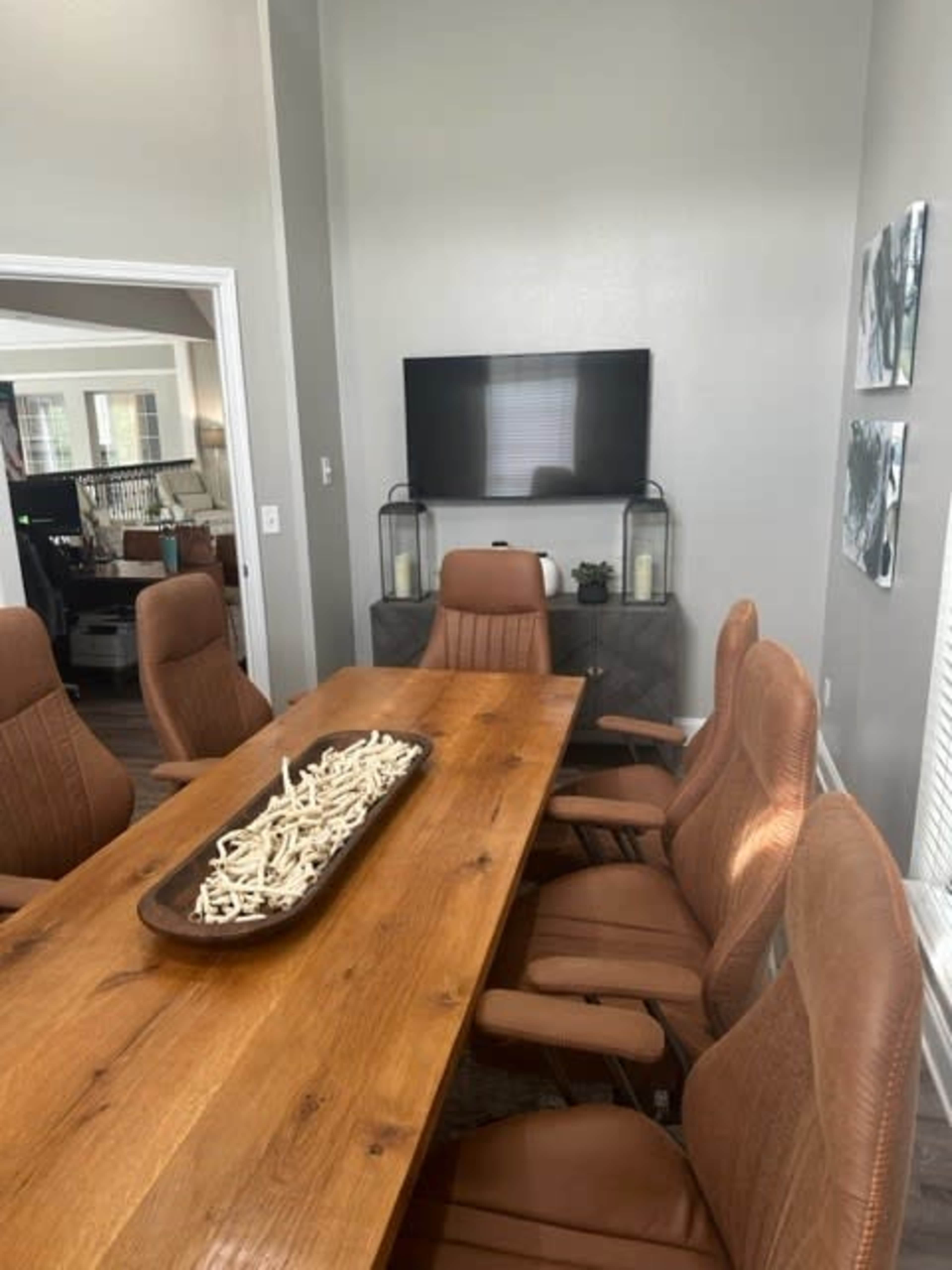 A modern conference room features a large wooden table surrounded by brown leather chairs, with a television mounted on the wall and decorative items displayed on a sideboard.