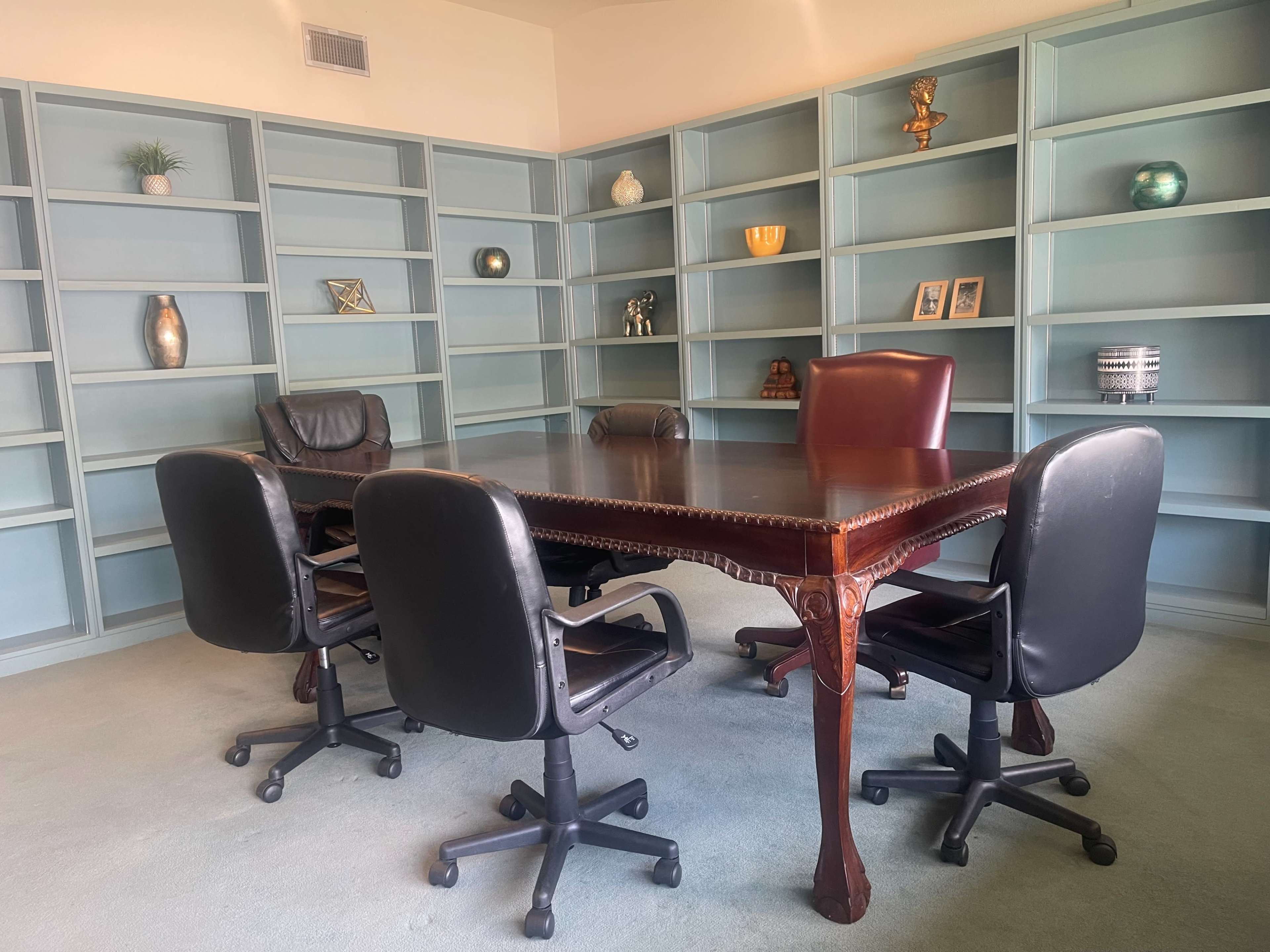 A conference room features a large wooden table surrounded by four black rolling chairs, with built-in shelves displaying various decorative items in the background.