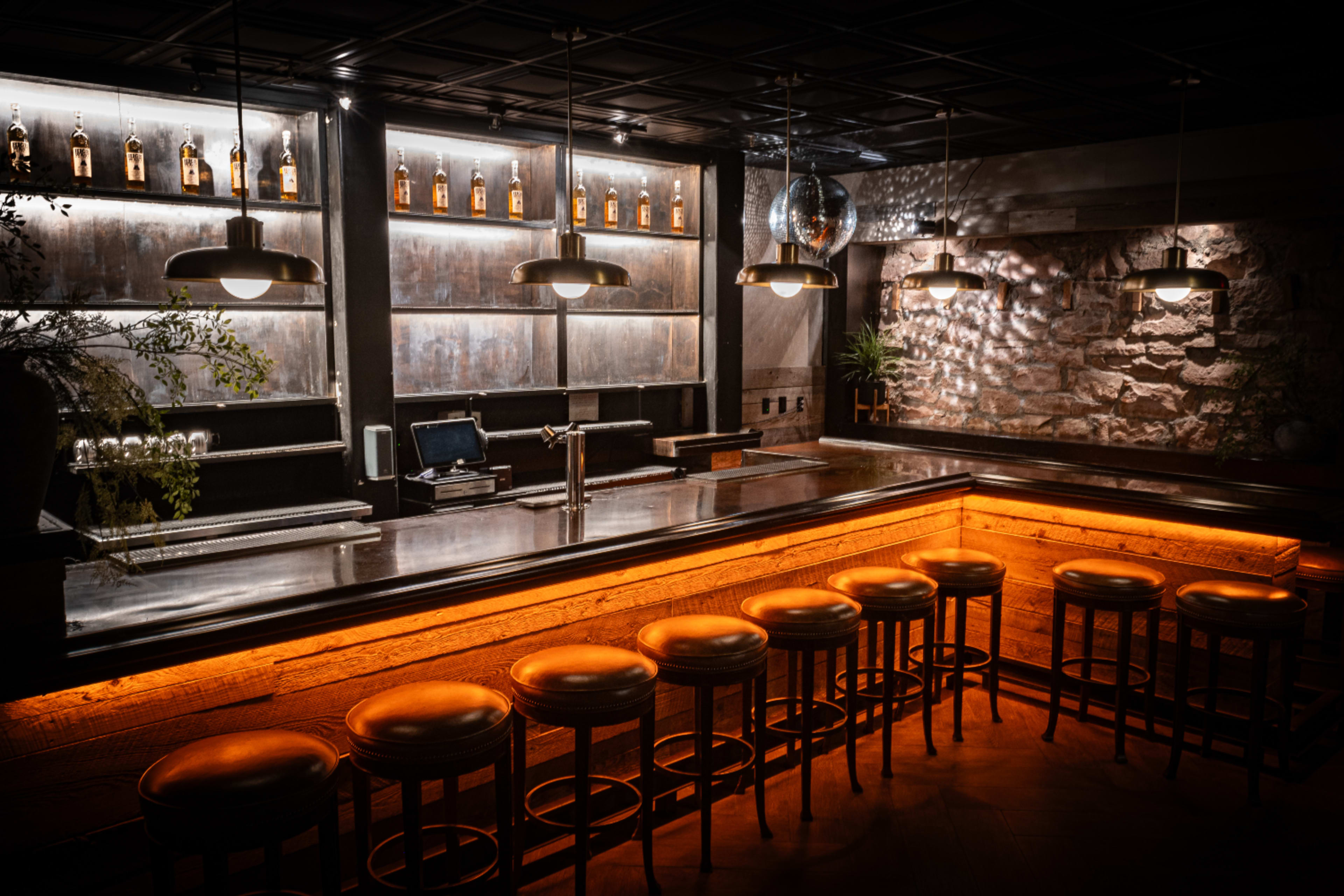 A dimly lit bar with a polished countertop, wooden stools, and shelves displaying bottles of liquor.