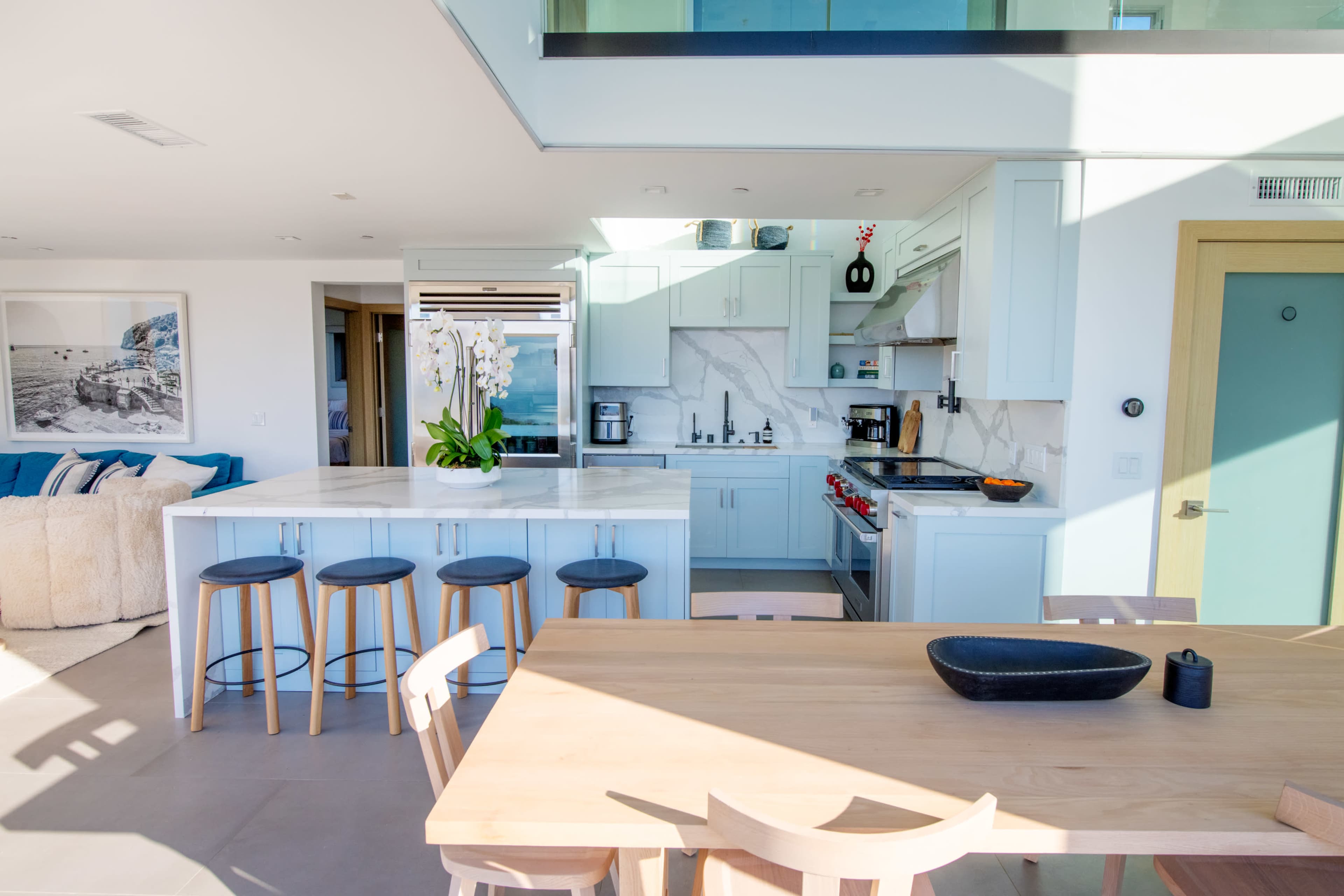A modern kitchen features light blue cabinetry, a marble countertop island with bar stools, and a wooden dining table.