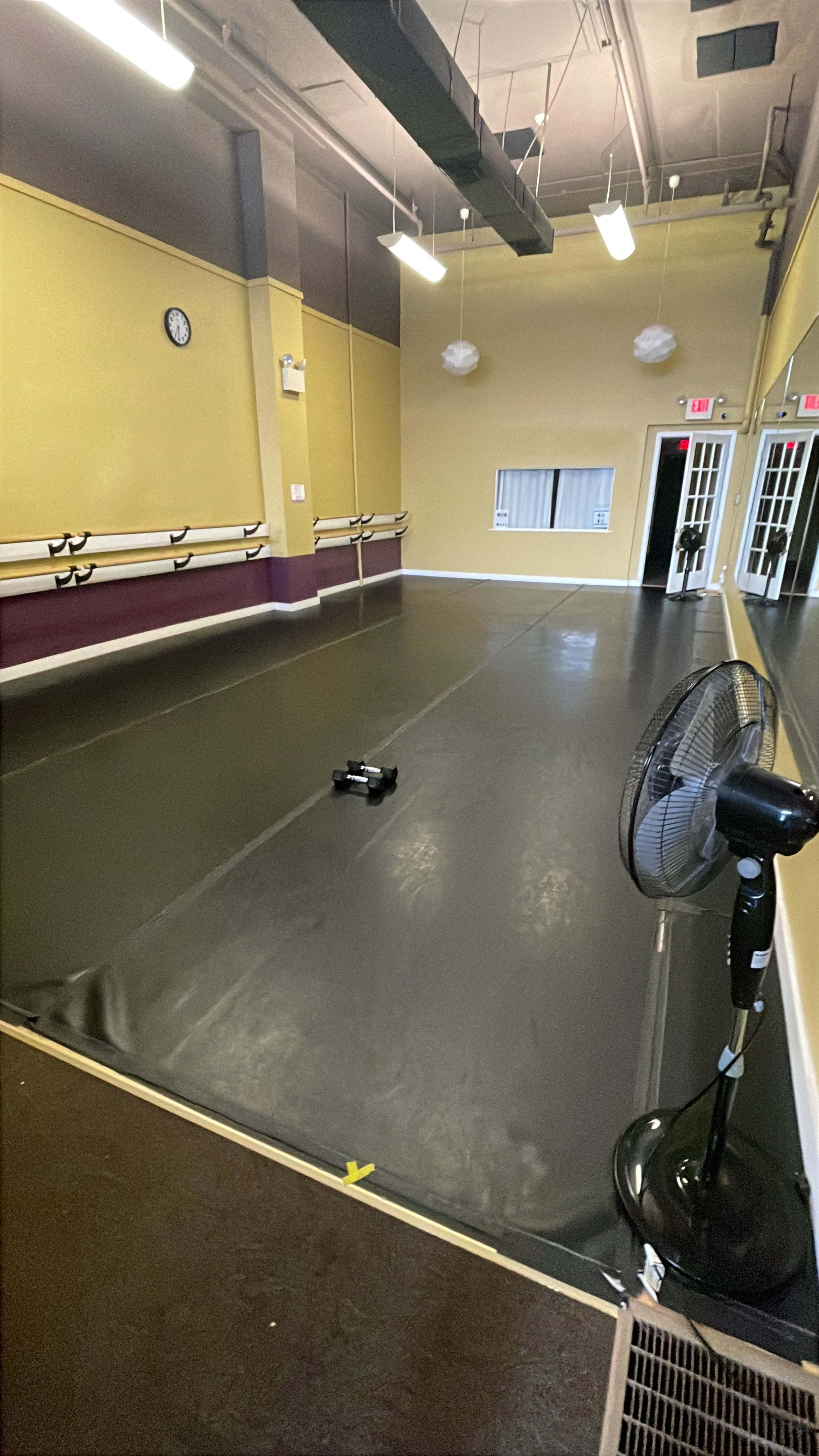 The image shows an empty dance studio with a smooth black floor, ballet bars along the walls, and a fan in the corner.