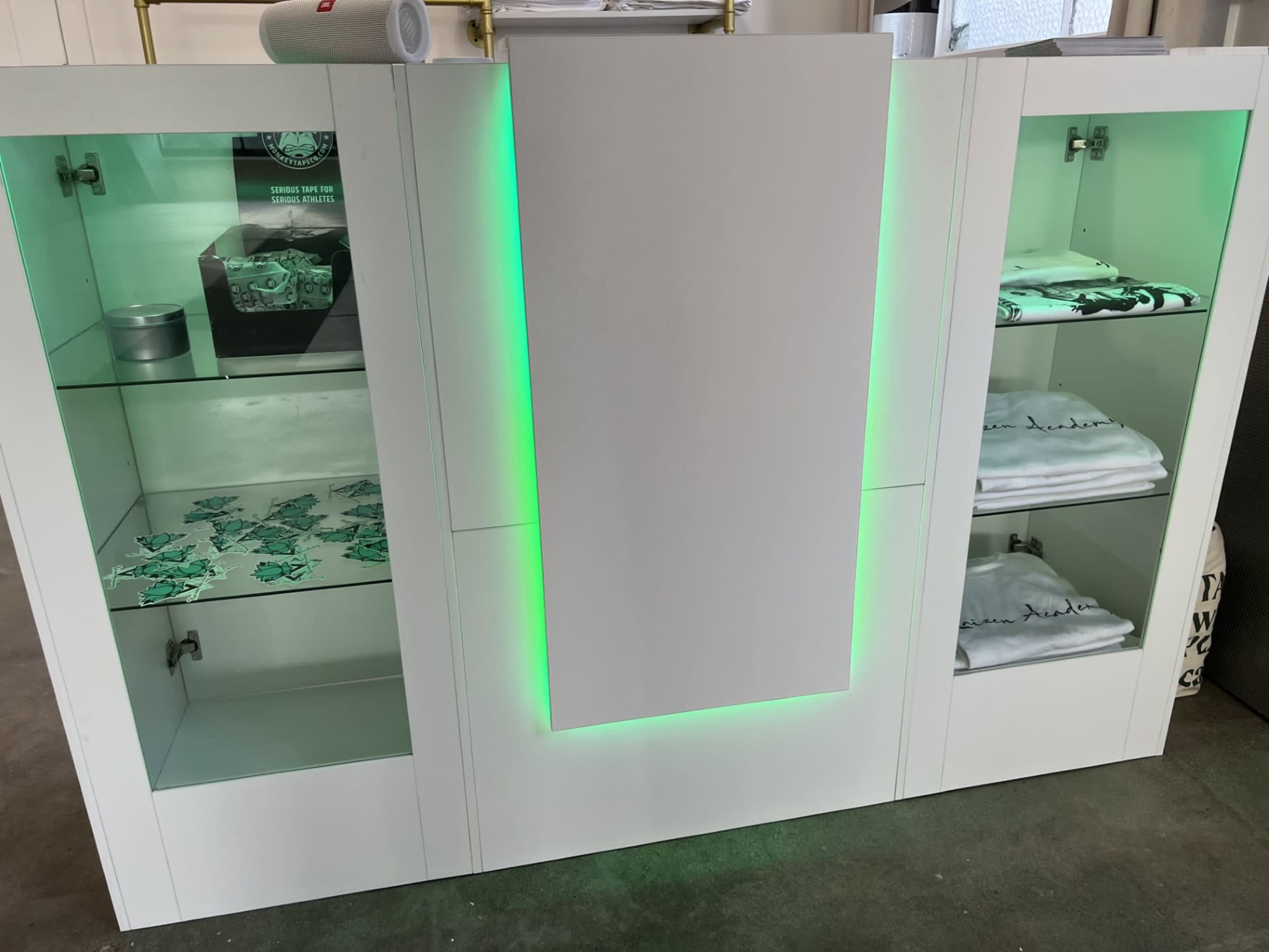 The image shows a modern display cabinet with illuminated green accents, featuring shelves that hold various items, including folded shirts and boxes.