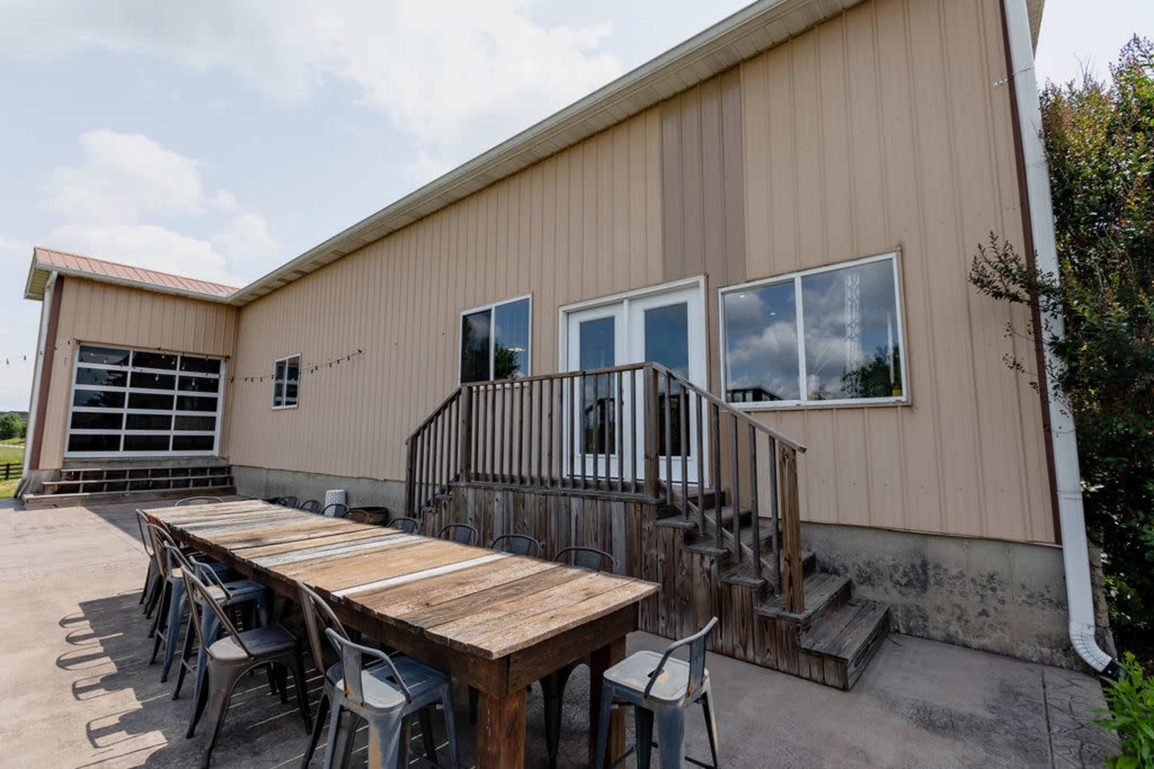 A long wooden table with metal chairs is set outside a large, light-colored building with a deck and stairs leading to a double door entrance.