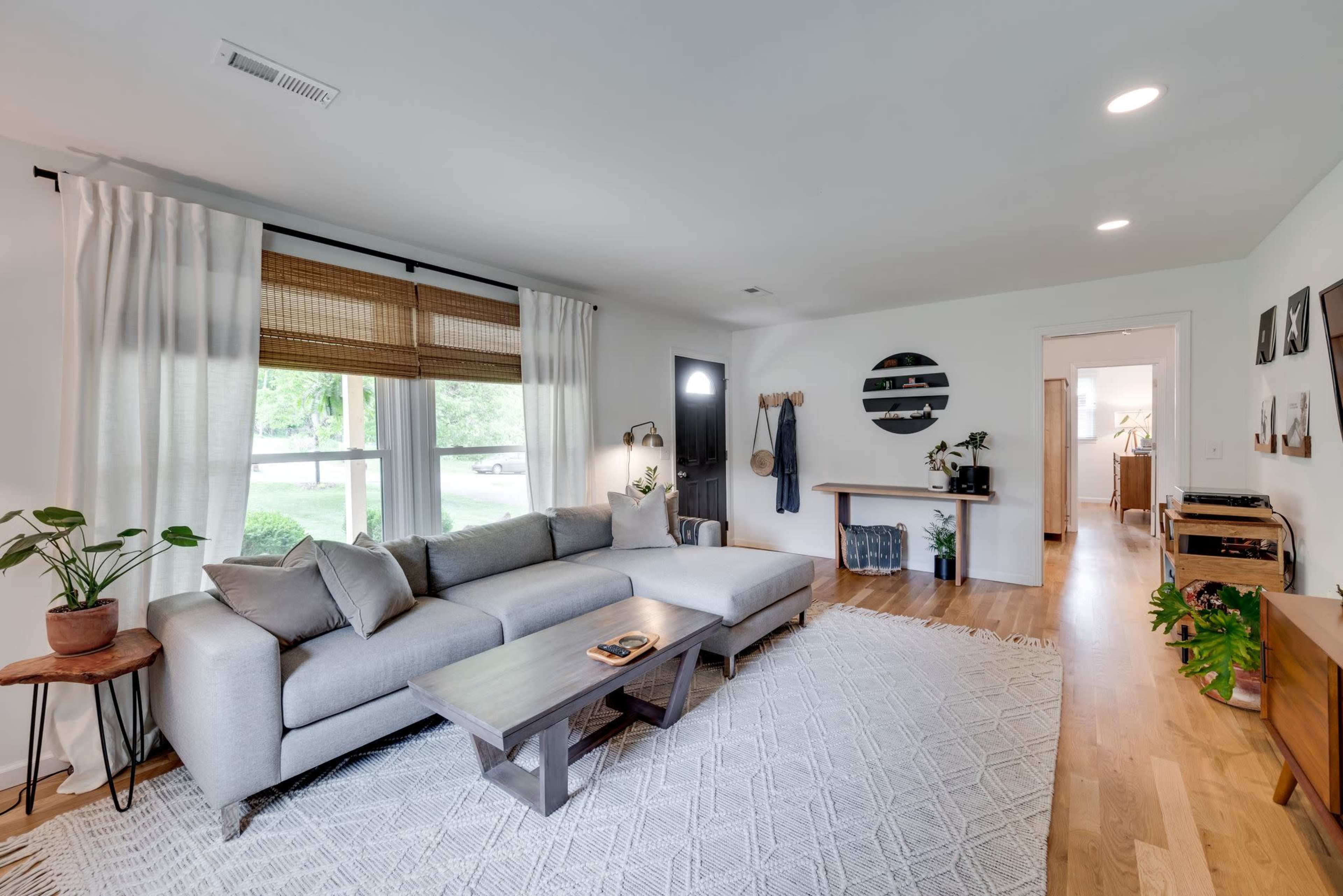 A bright living room featuring a gray sectional sofa, a wooden coffee table, and plants, with a visible entryway leading to a kitchen area.