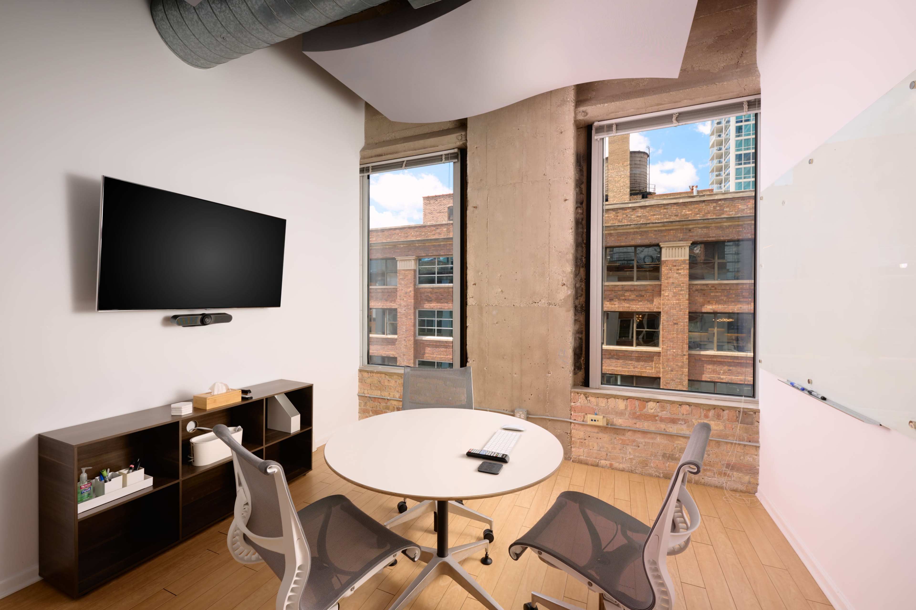 A modern meeting room features a round table, chairs, a large wall-mounted TV, and windows overlooking brick buildings.