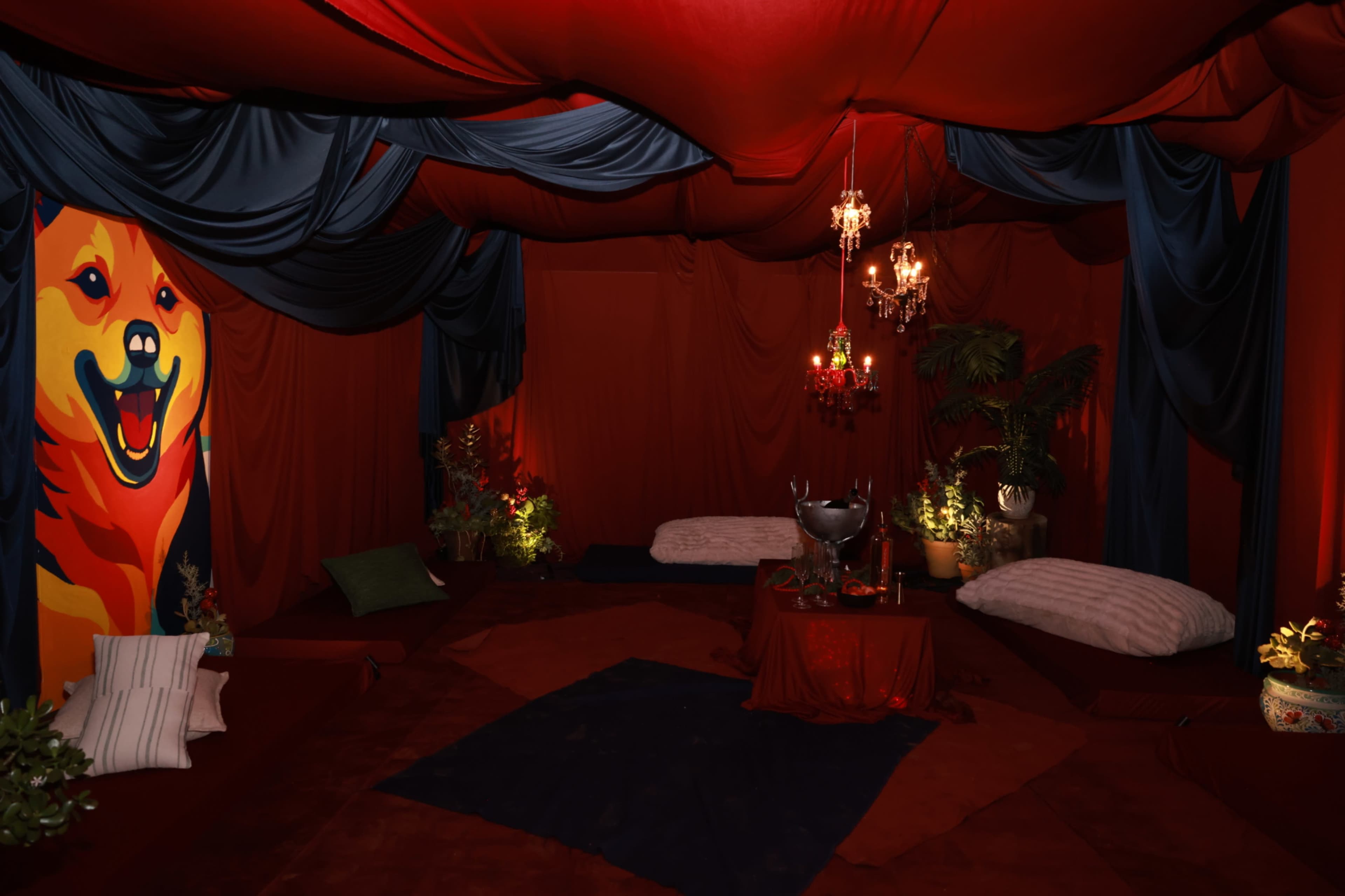 The image depicts a dimly lit room draped in red and blue fabric with a colorful mural of a dog on one wall, featuring plush seating and decorative plants.