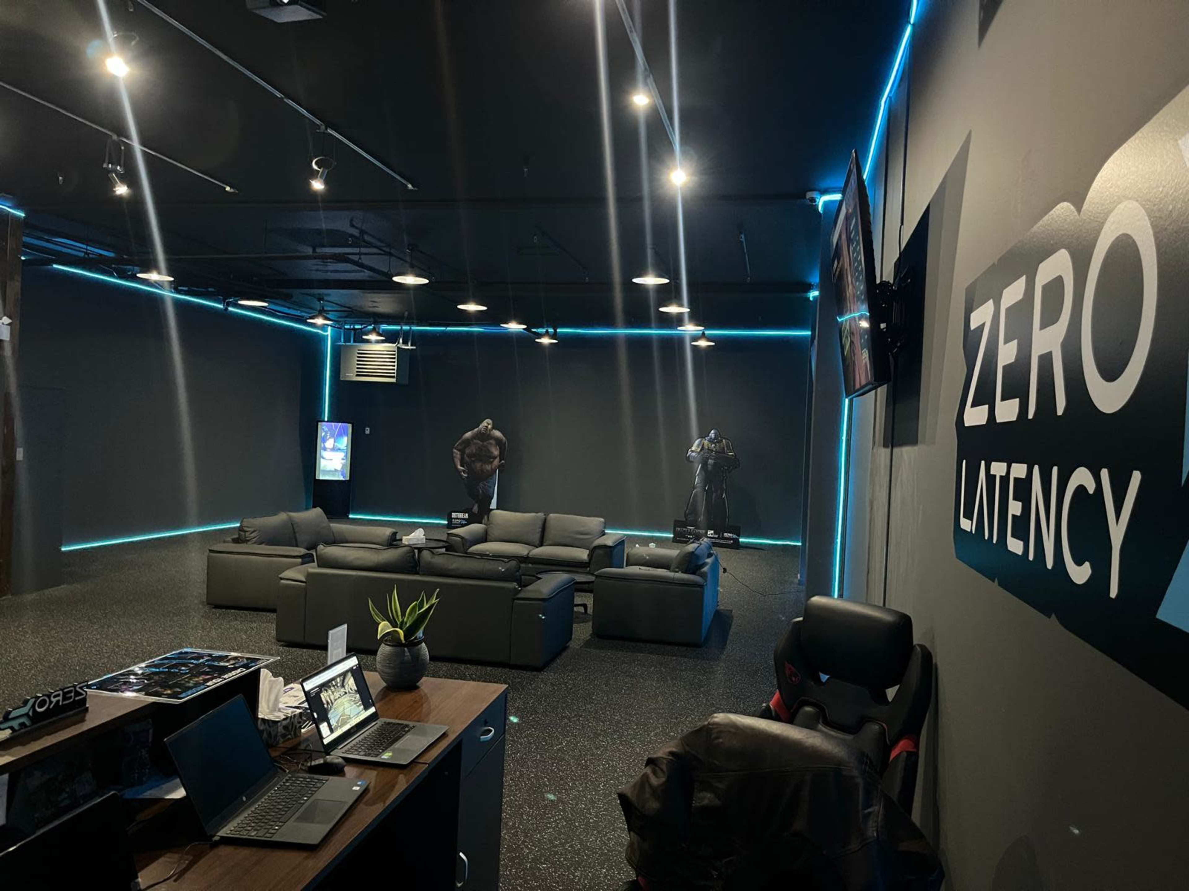 The image shows a gaming lounge with gray walls, a large sectional sofa, and neon blue lighting, featuring a desk with a laptop and a statue in the background.