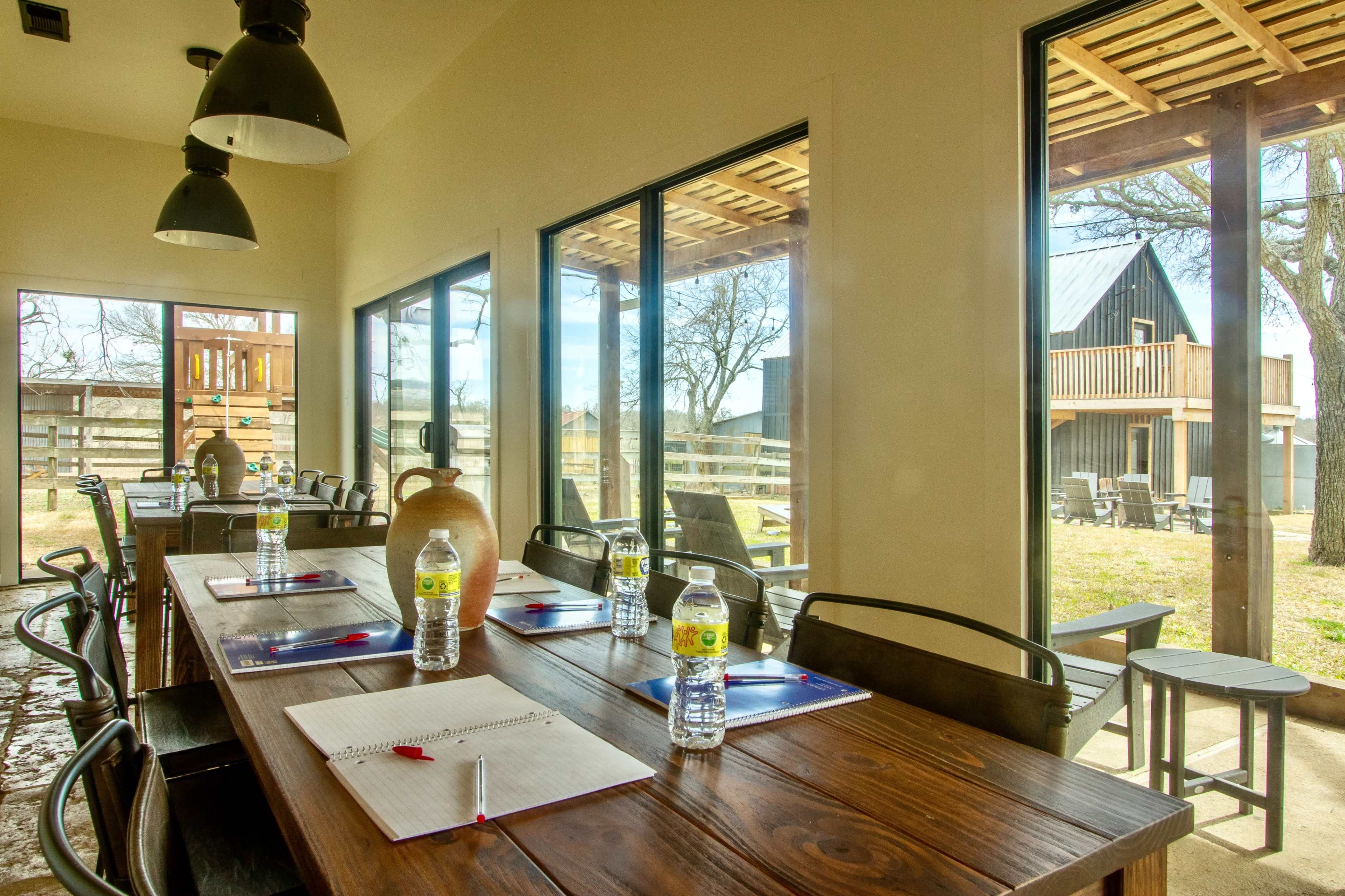 A long wooden table set with notebooks and water bottles is arranged in a brightly lit meeting room with large windows overlooking an outdoor area.