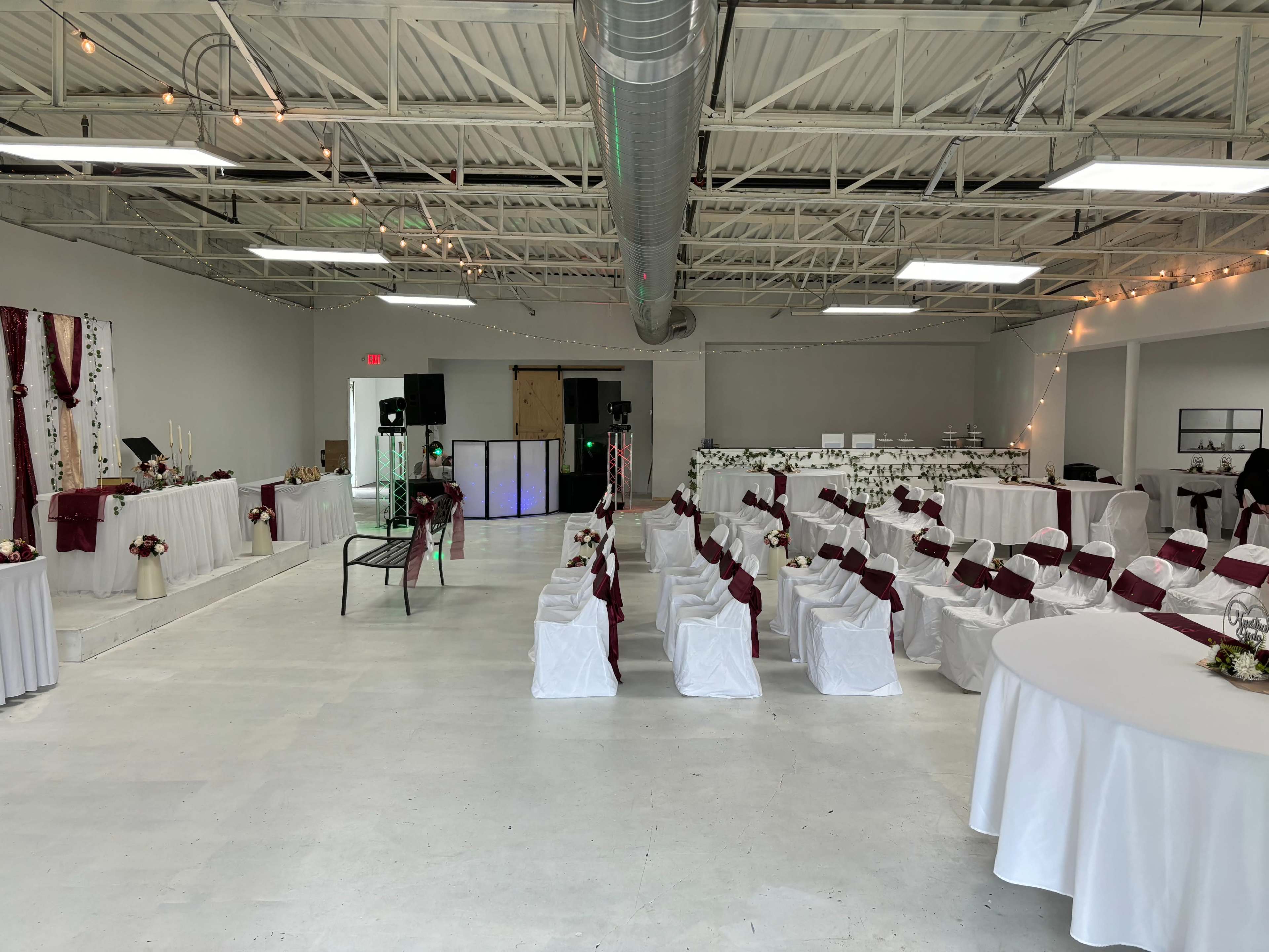 A spacious event venue set up for a ceremony, featuring rows of white chairs with maroon sashes, a decorated stage area, and tables covered with white cloths.