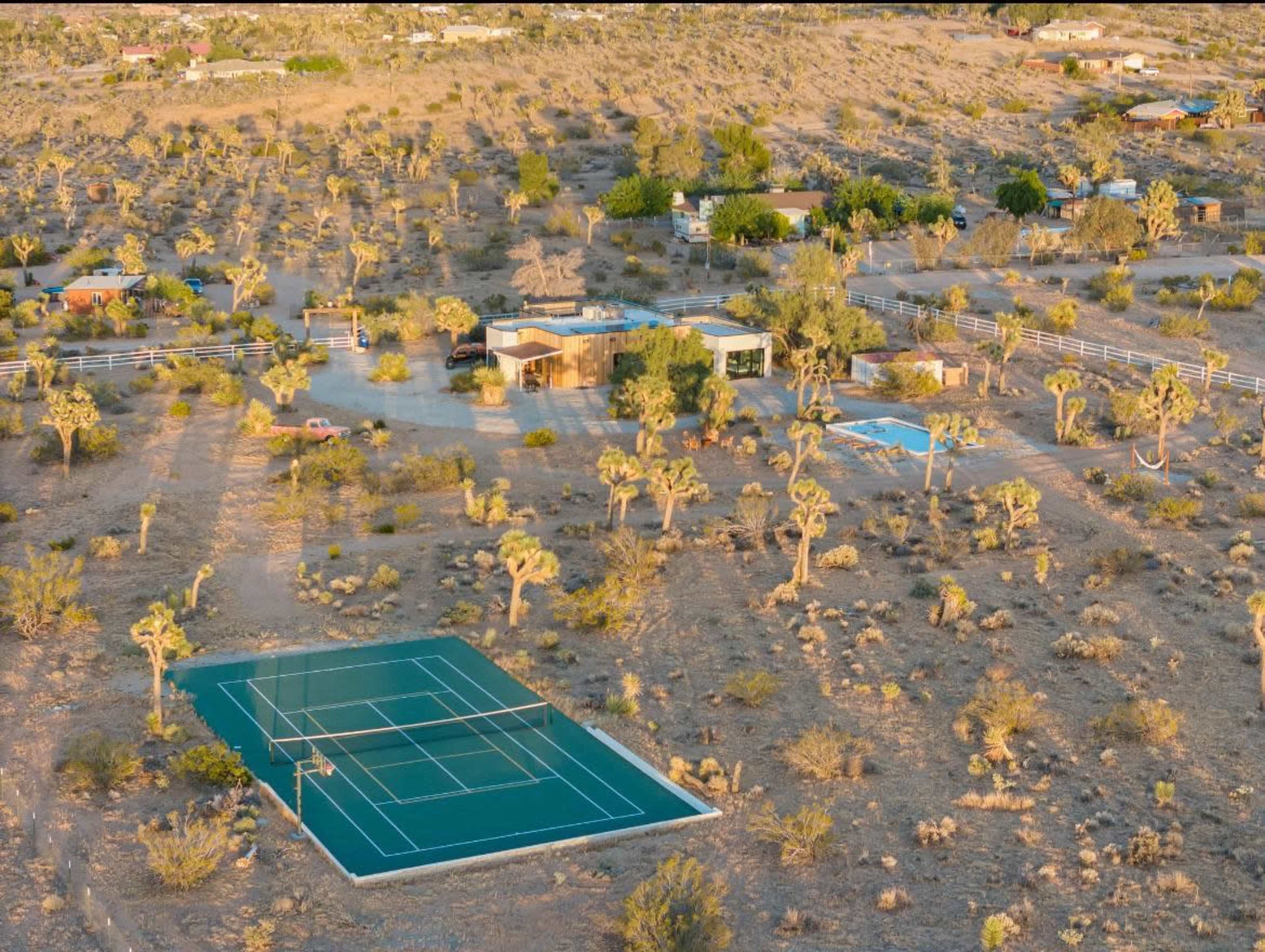 A desert landscape featuring a tennis court and several structures surrounded by Joshua trees and sparse vegetation.