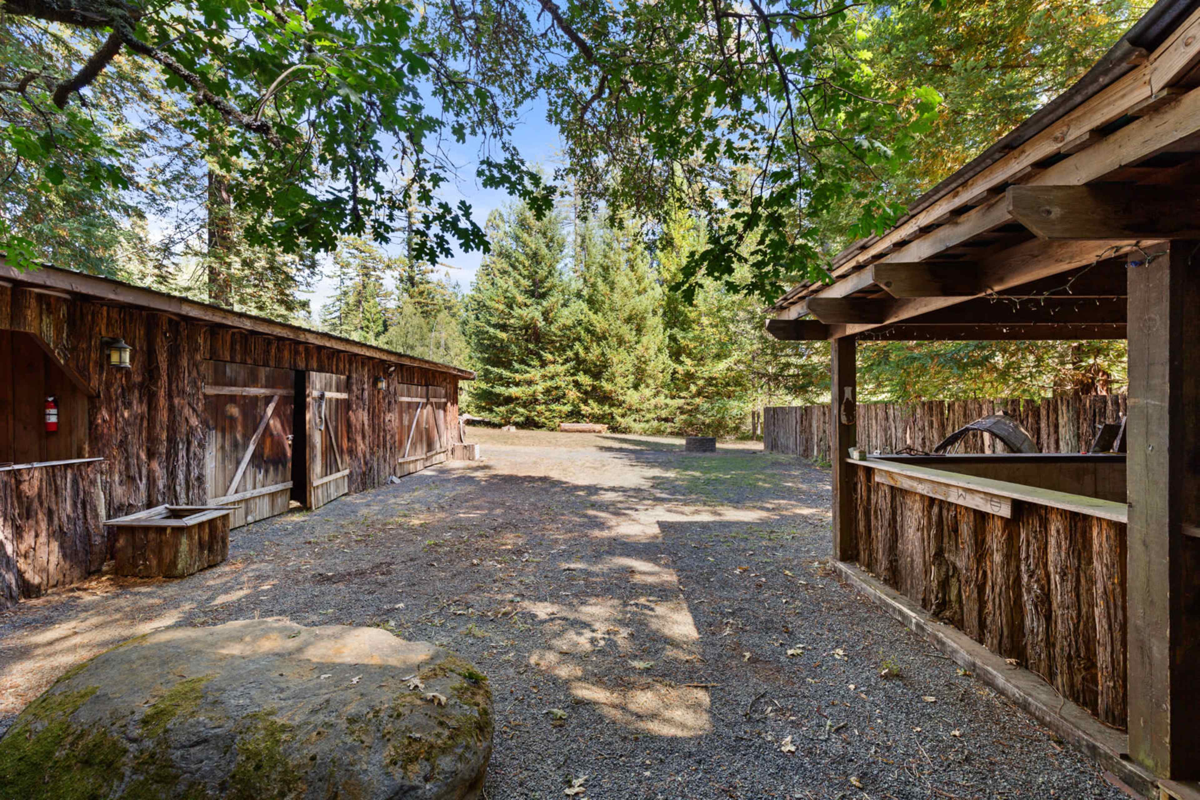 The image shows a rustic outdoor area featuring two wooden structures, a large rock, and surrounding trees under a clear sky.