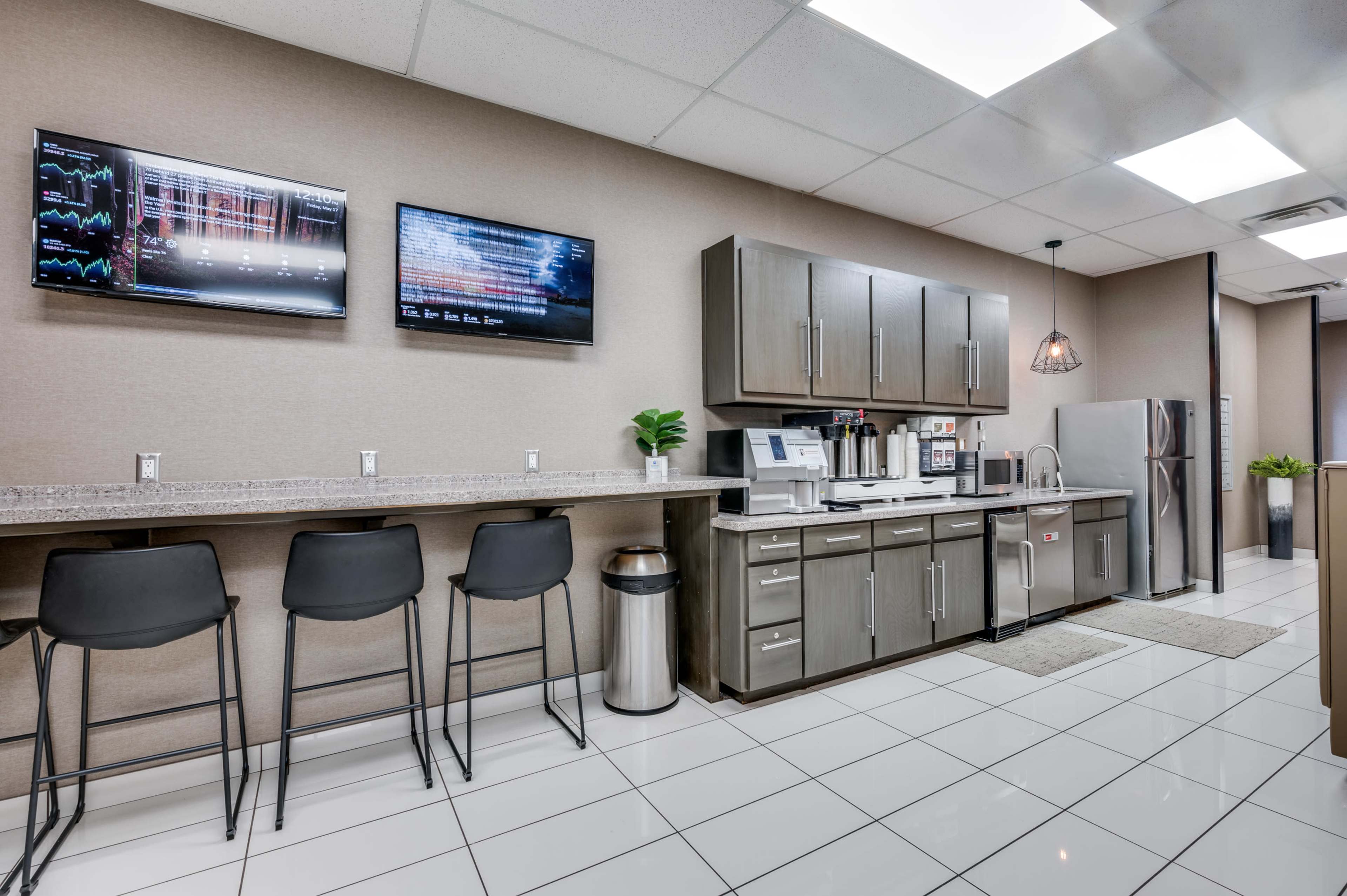 The image shows a modern break room featuring a kitchenette with two television screens mounted on the wall, a counter with barstool seating, and appliances including a refrigerator and coffee maker.