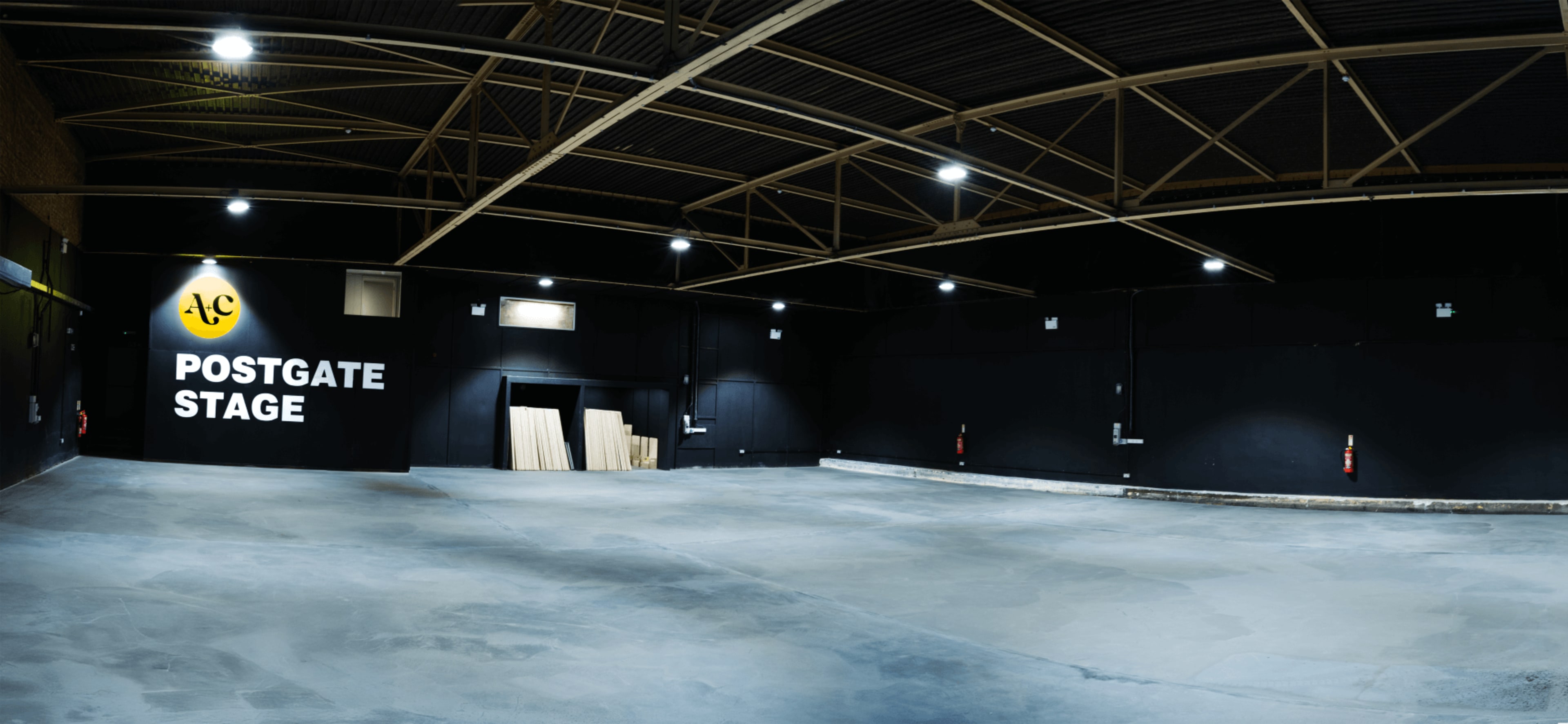 The image shows an empty, well-lit warehouse space with black walls and a sign reading "POSTGATE STAGE."