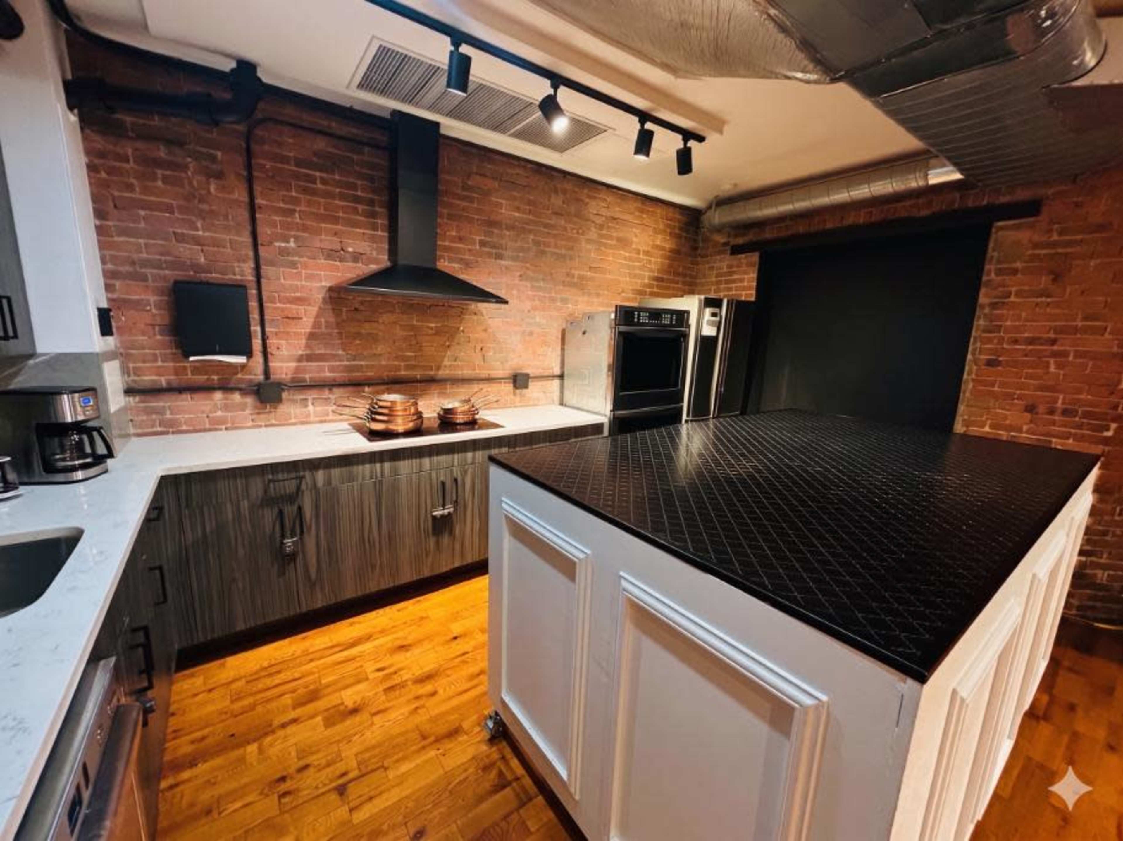 The image shows a modern kitchen featuring exposed brick walls, a central black countertop island, stainless steel appliances, and dark cabinetry.