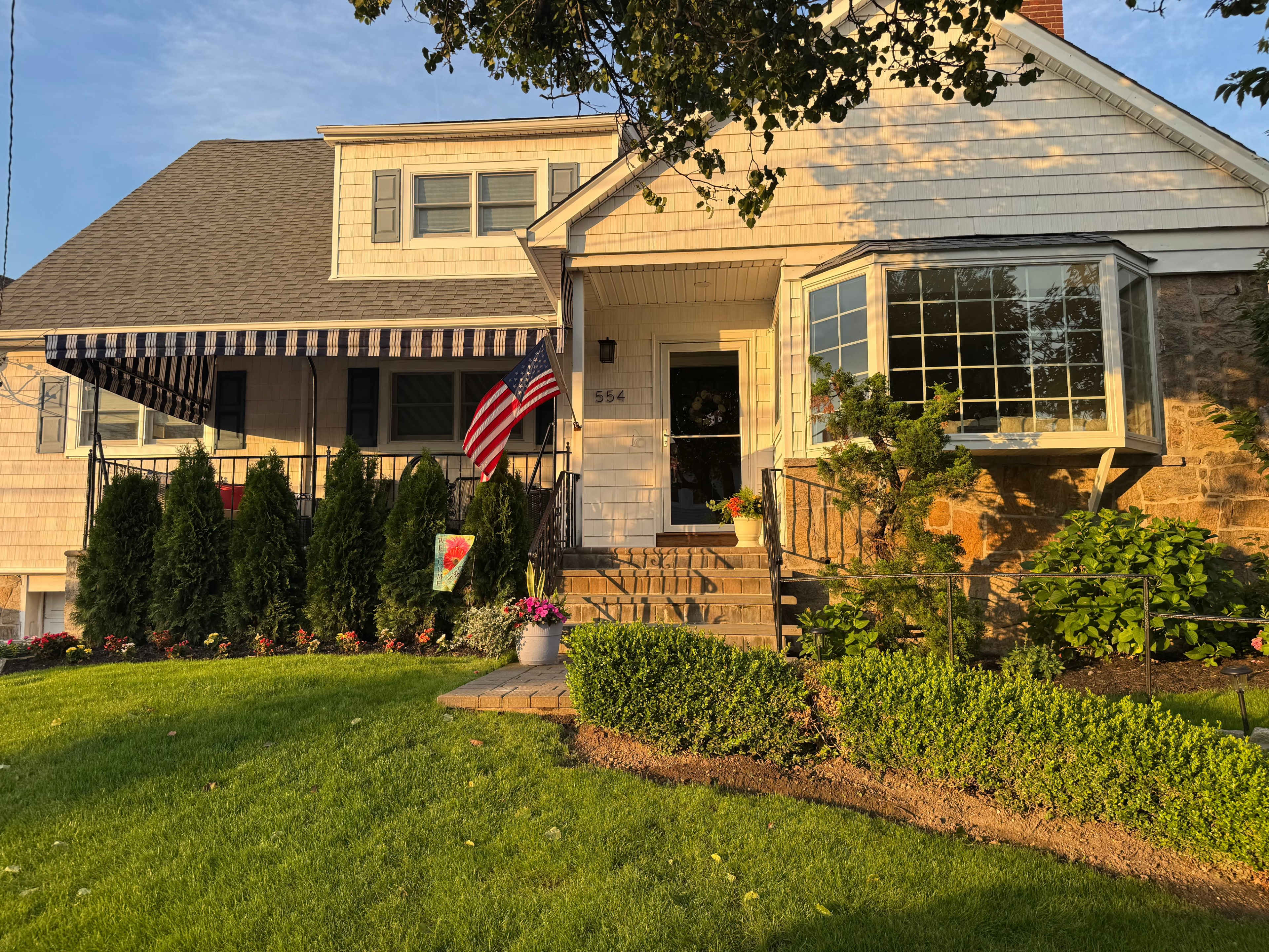 A house features a stone facade with a welcoming porch, surrounded by neatly maintained landscaping and an American flag displayed.