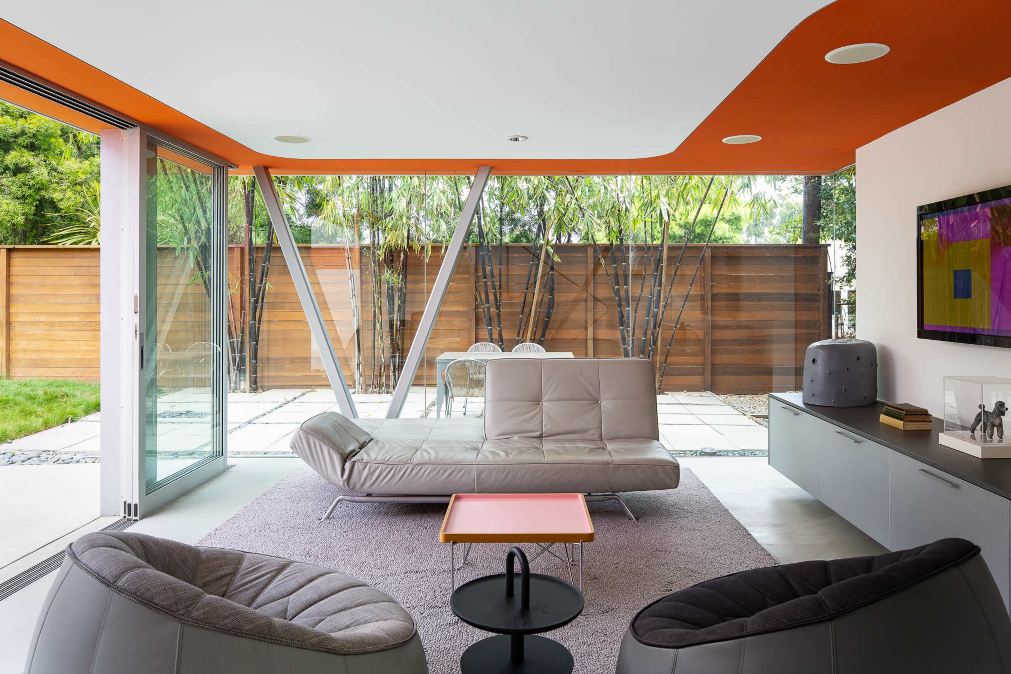 A modern living room with a gray sectional sofa, two black lounge chairs, and floor-to-ceiling windows that overlook a landscaped backyard with wooden fencing.