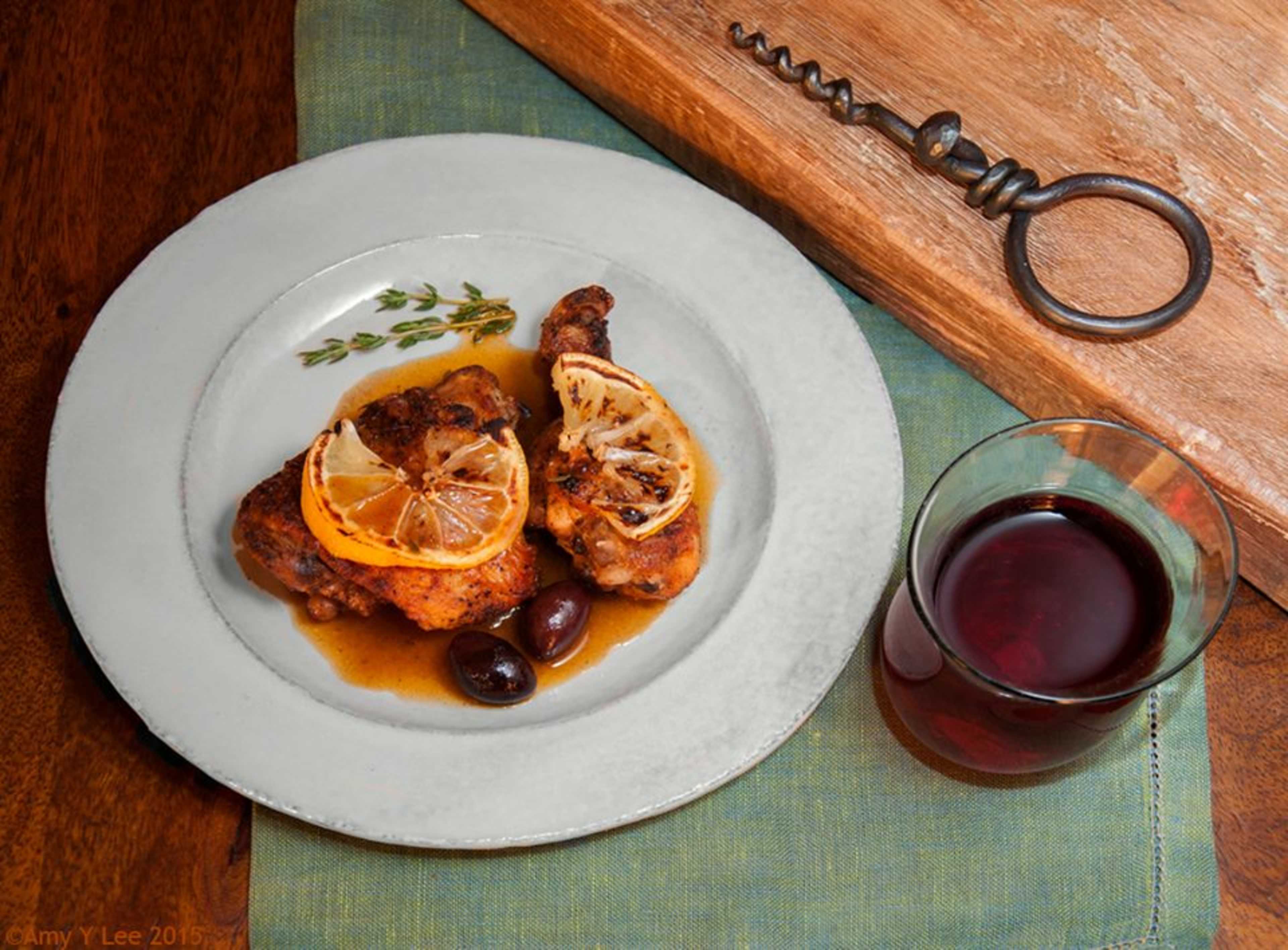 The image shows a plate of chicken drumsticks garnished with lemon slices and olives, accompanied by a glass of red wine on a wooden table.