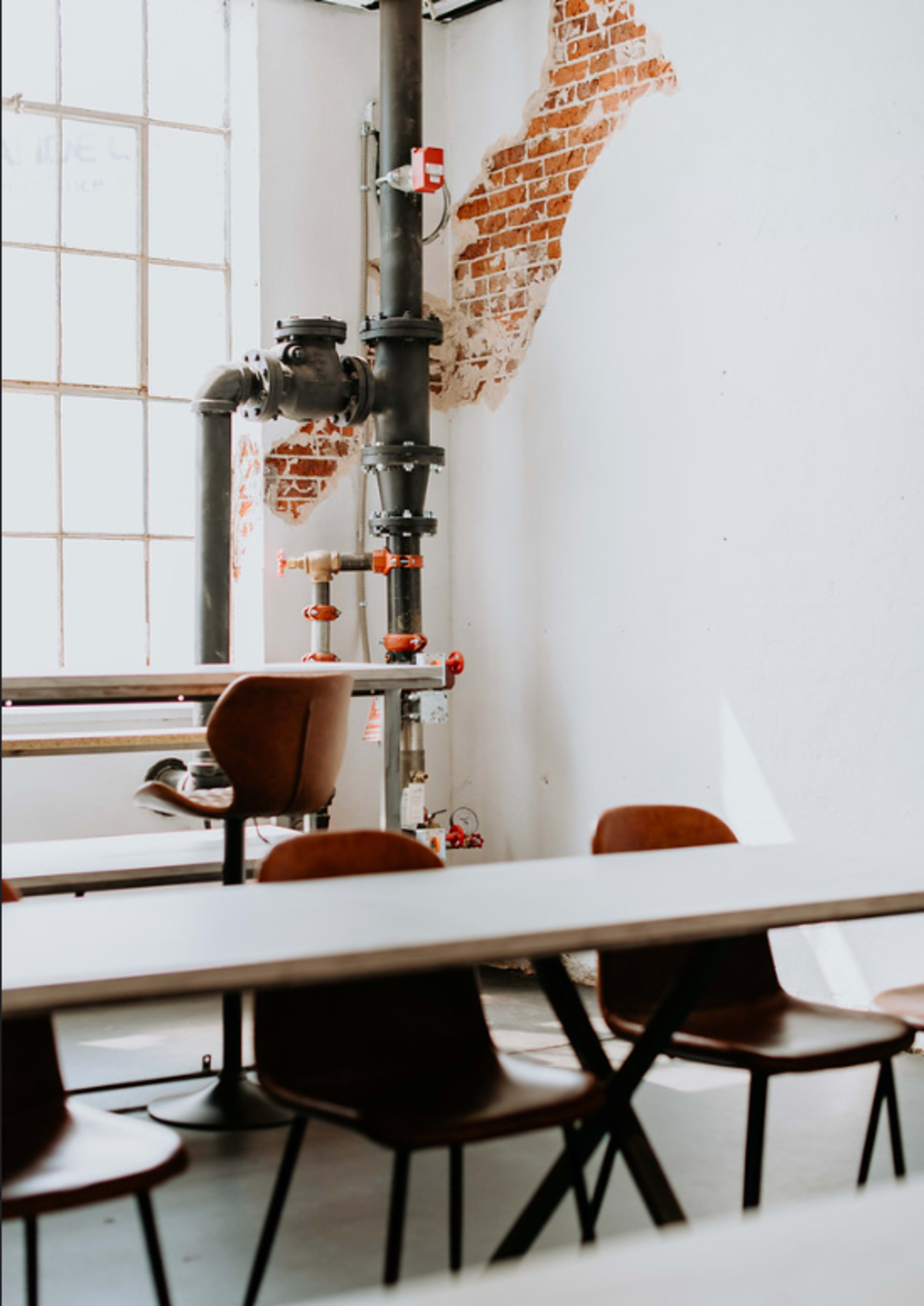 A bright interior features a series of simple tables and chairs along with exposed brick and industrial pipes on the wall.