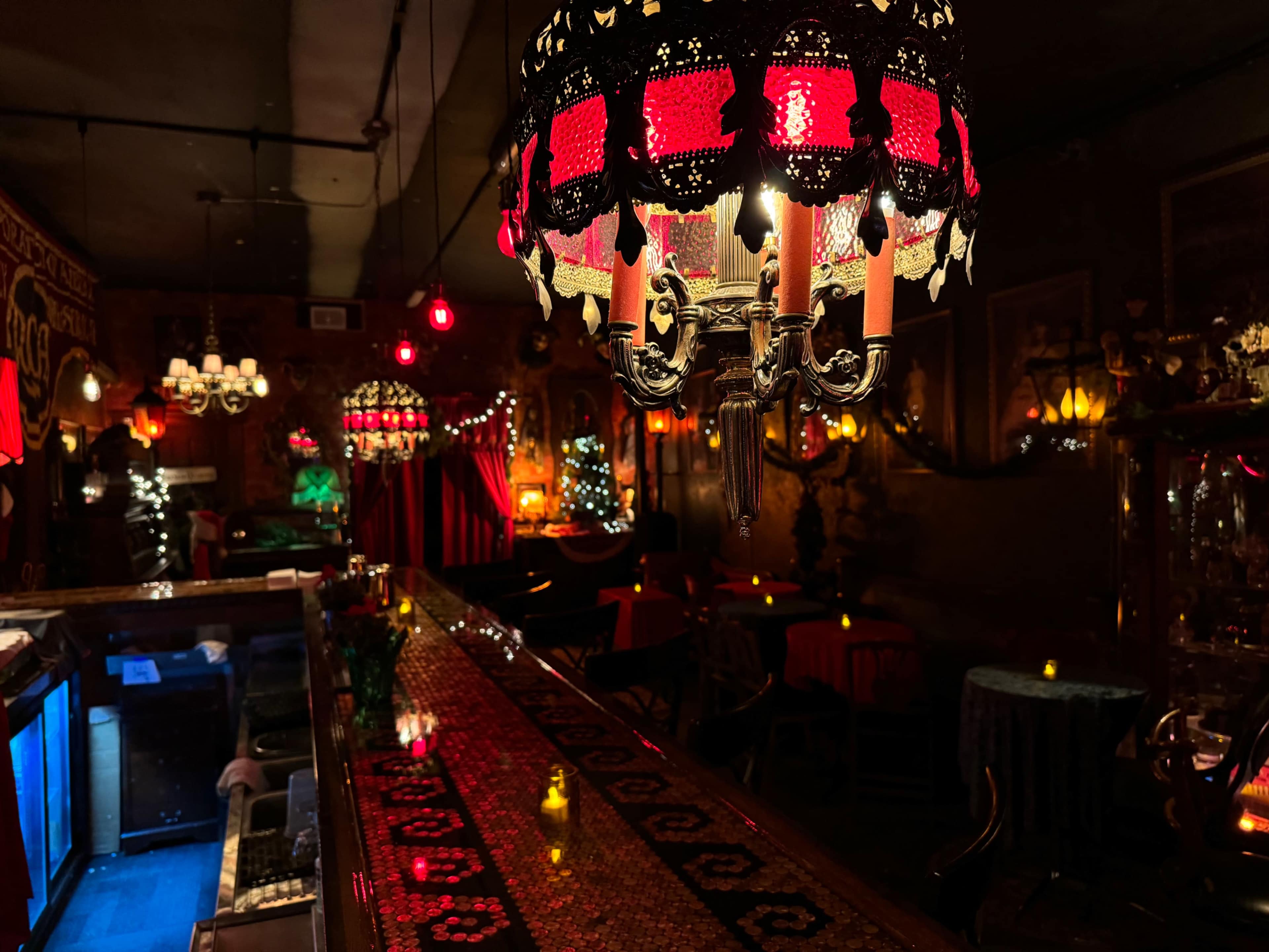 A dimly lit bar with ornate red chandeliers, a long wooden counter, and small tables adorned with candles.