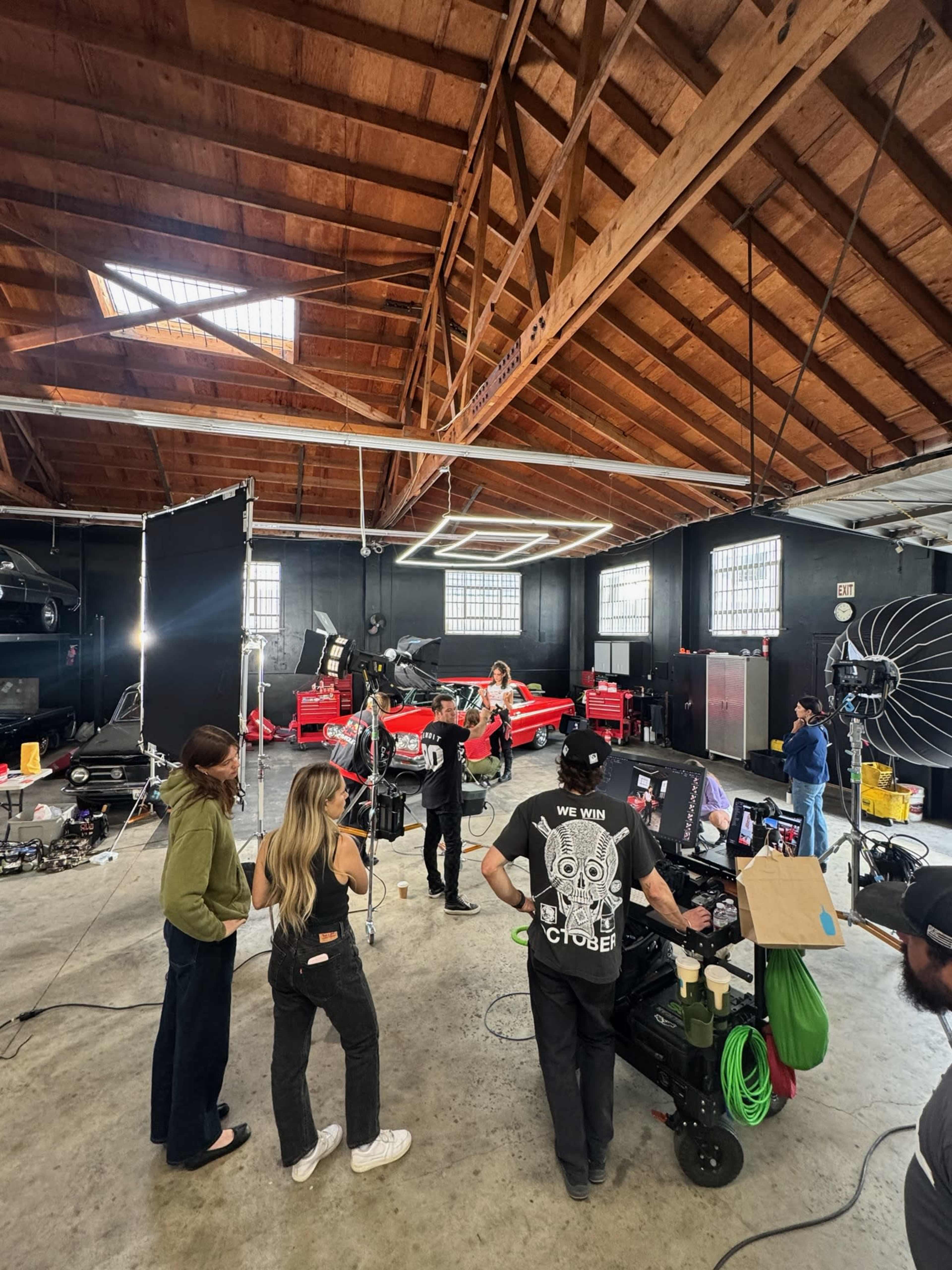 A group of people is working on a film or photo shoot in a spacious studio, with a red car positioned in the center under bright lights and equipment.