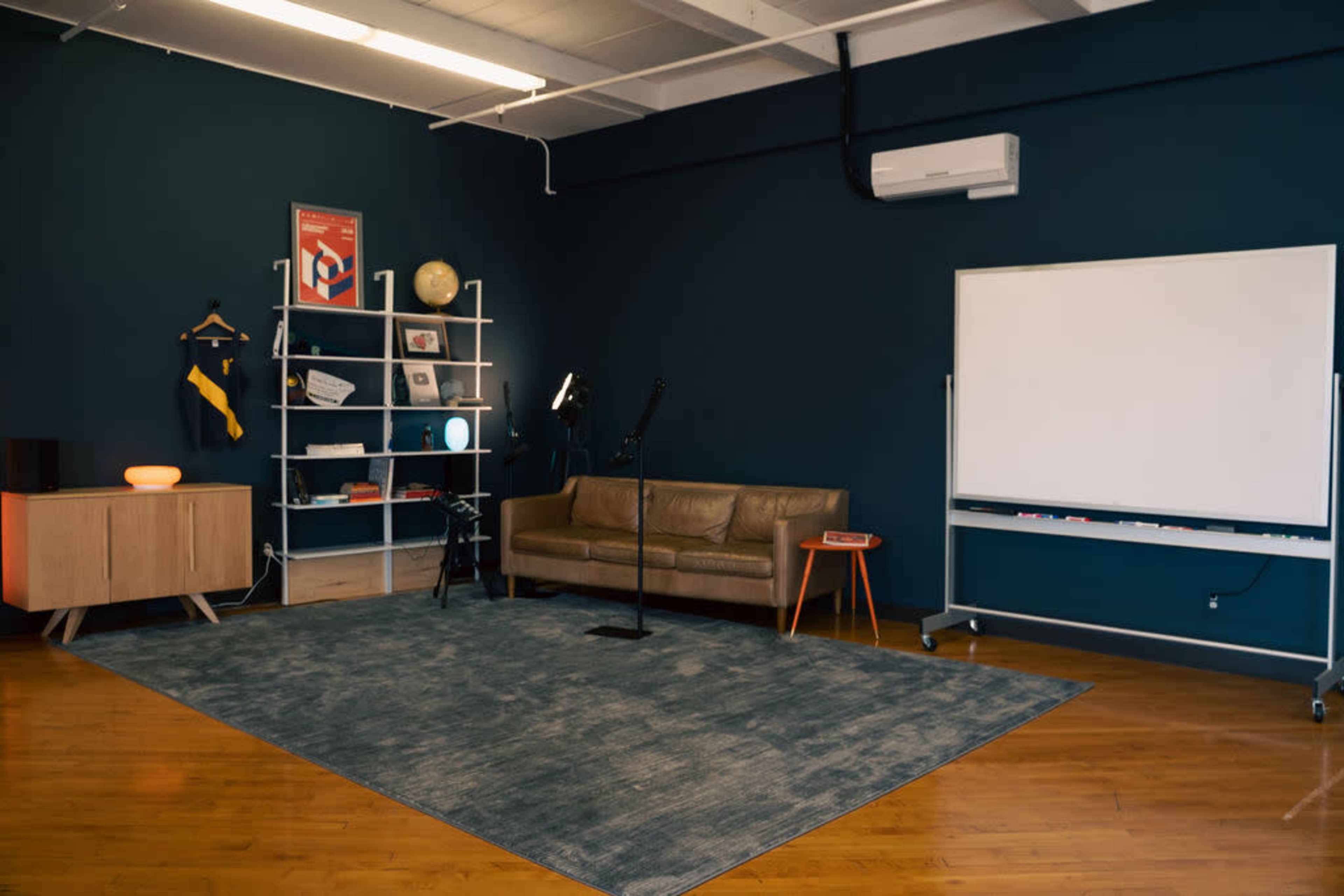 The image shows a modern, minimalist room with a couch, a bookshelf, a whiteboard, and a microphone stand on a large area rug.