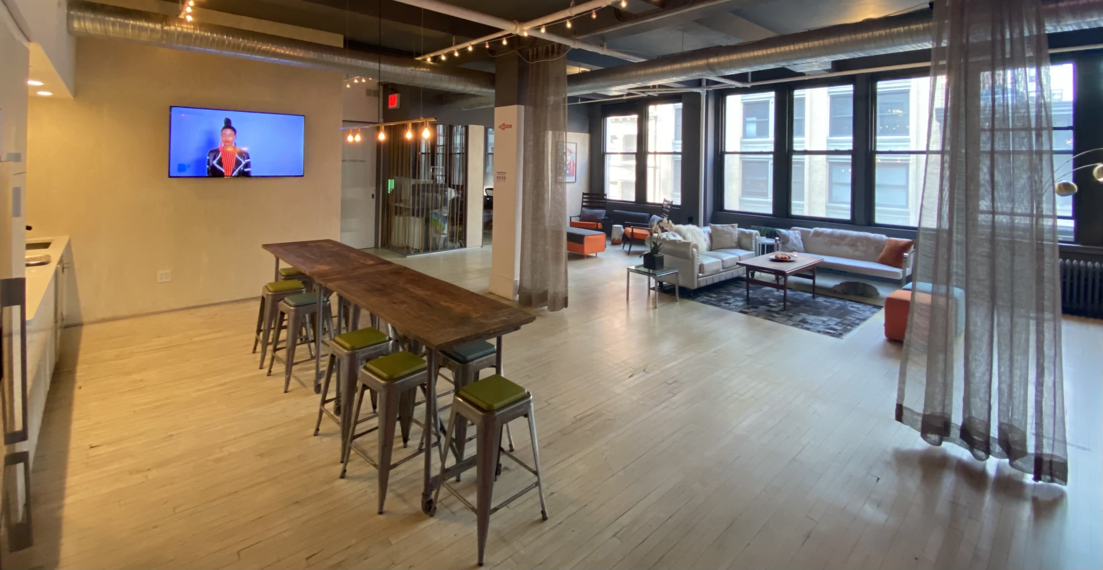 A spacious, modern open-plan living area with a long wooden table, metal stools, a comfortable seating arrangement, and a TV mounted on the wall.