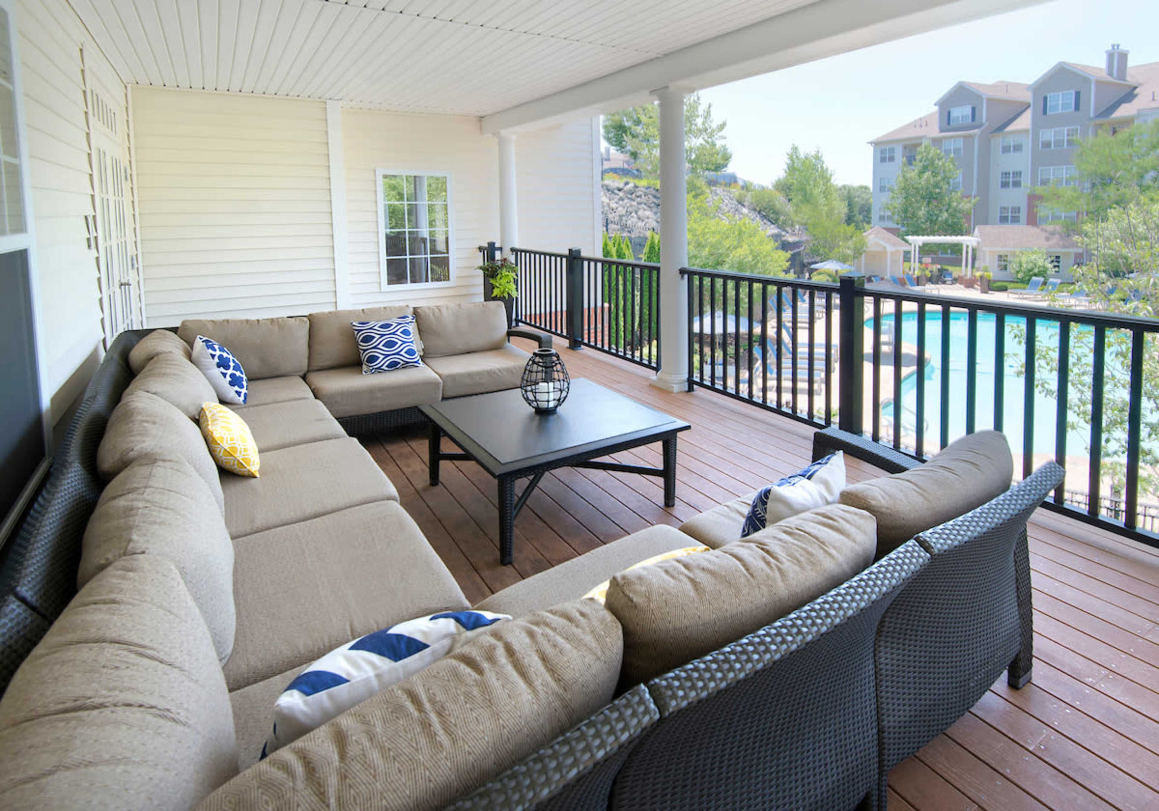 A spacious outdoor lounge area features a large sectional sofa arranged around a central table, overlooking a swimming pool and resort-style buildings.