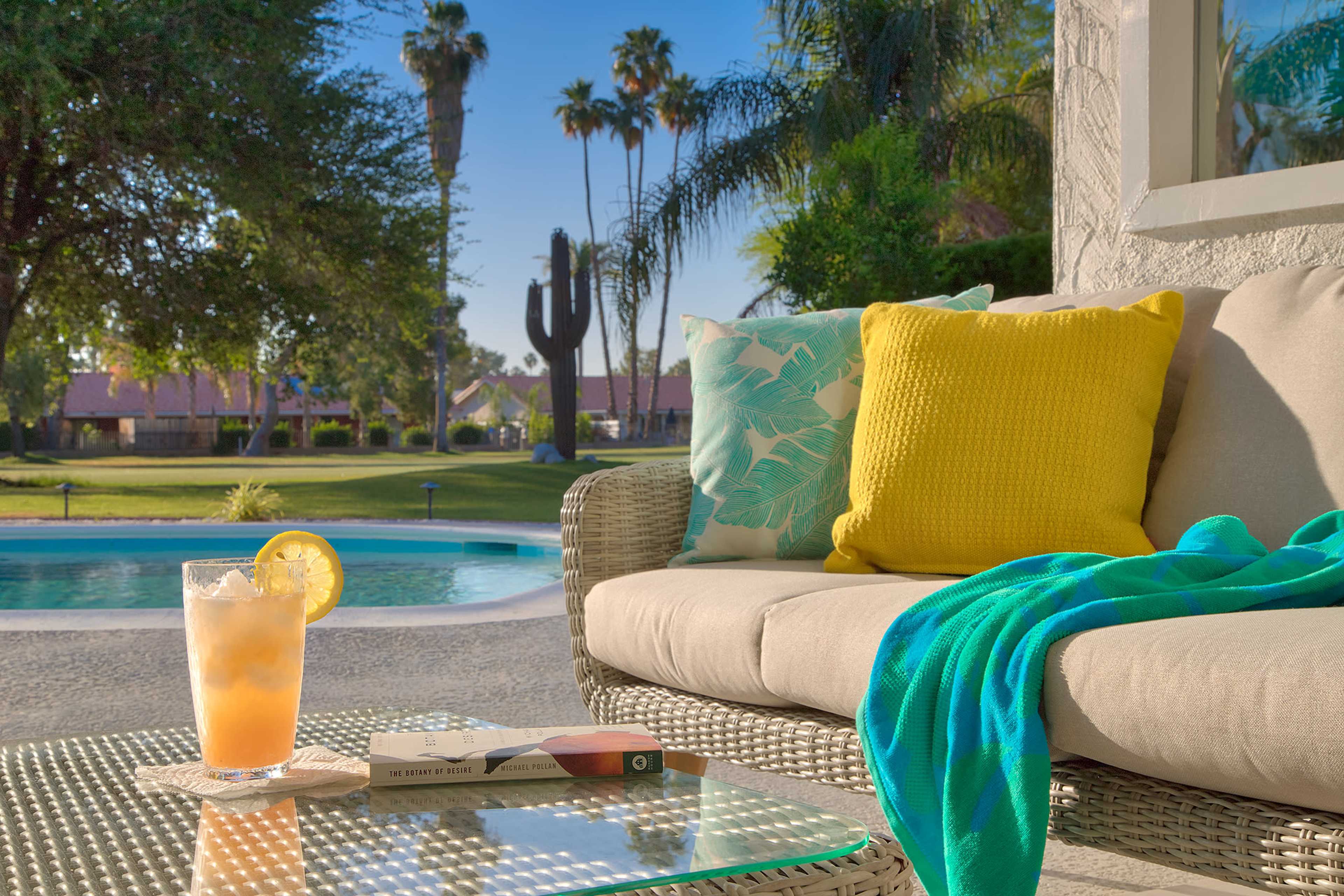 A cozy outdoor seating area features a glass of beverage with a lemon slice, surrounded by decorative pillows and a turquoise throw, overlooking a swimming pool and palm trees.