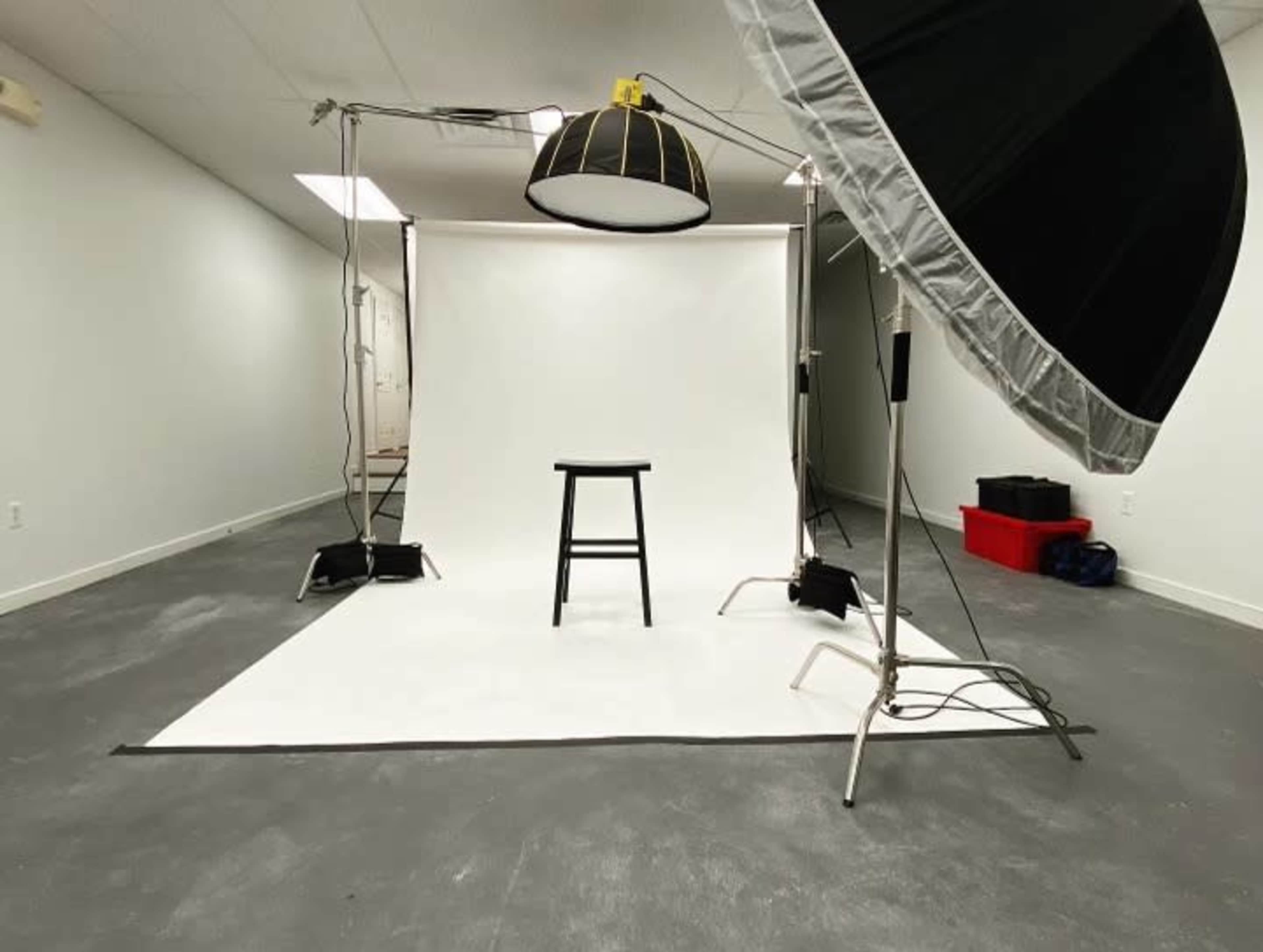 A minimalist photography studio featuring a black stool positioned on a white backdrop, with studio lighting equipment set up around it.