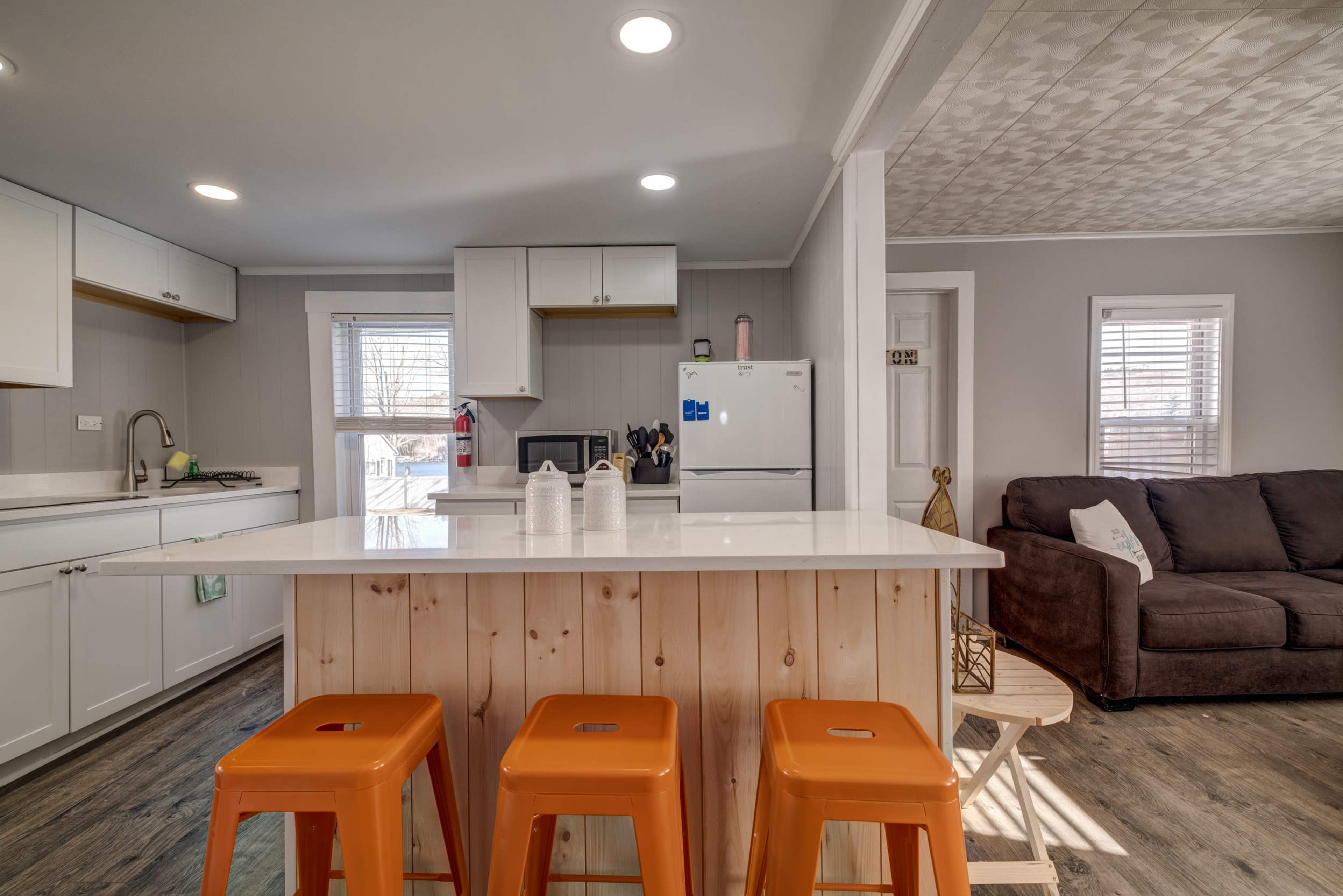 A modern kitchen with white cabinets, a central island with three orange stools, and a living area featuring a brown couch in the background.