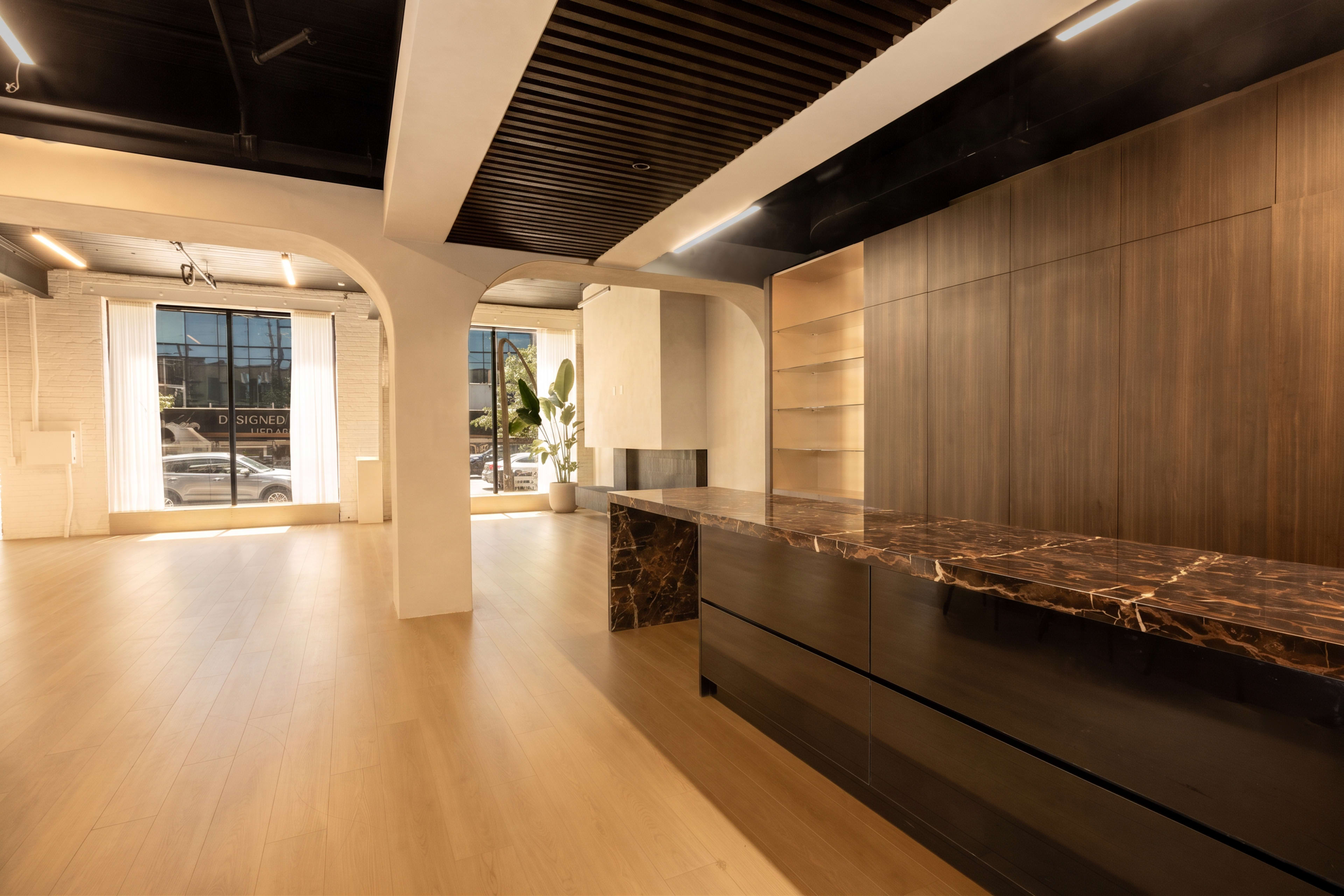 The image shows a modern interior space with a polished reception desk made of dark materials, large windows allowing natural light, and minimalistic decor.