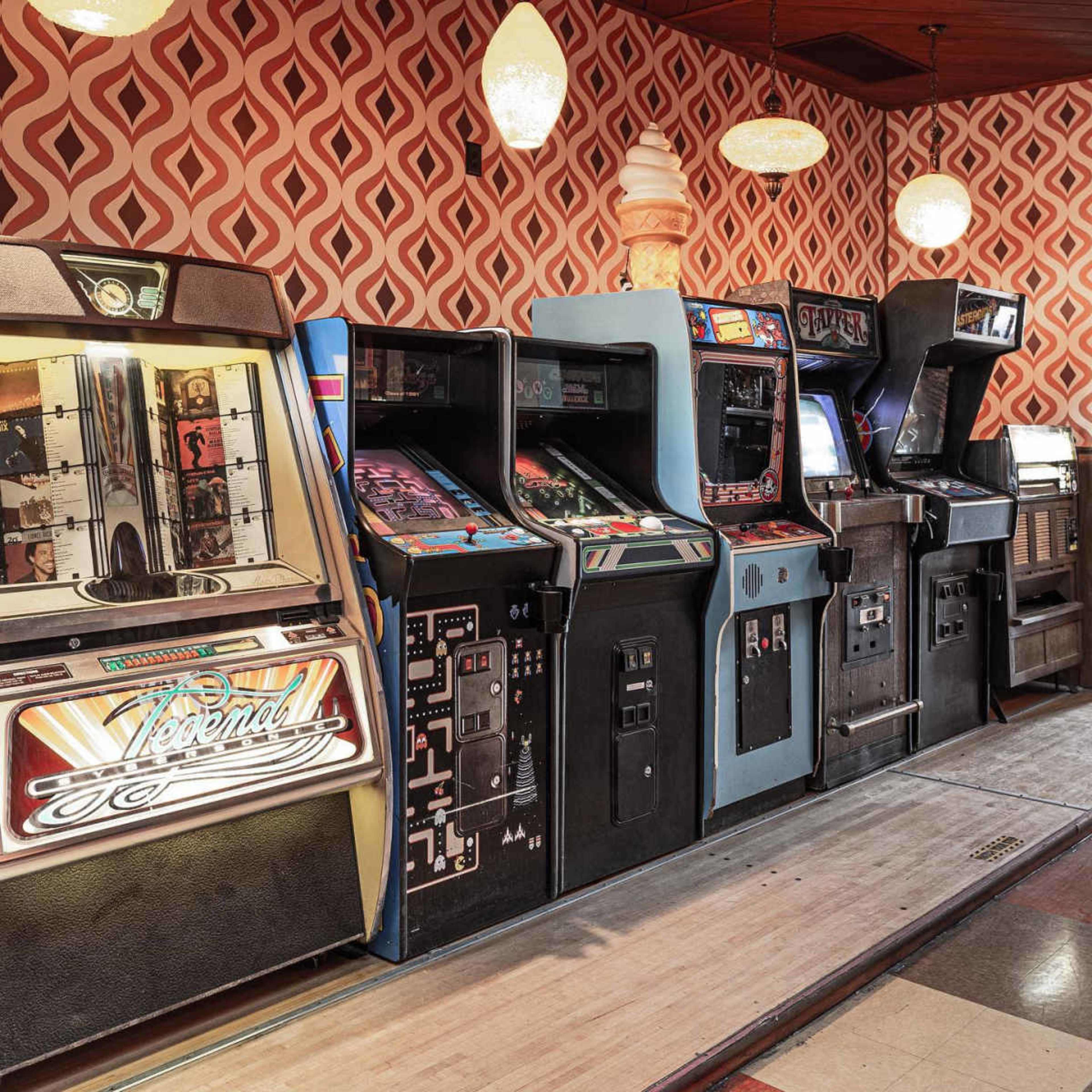The image shows a row of vintage arcade game machines against a patterned wallpaper backdrop in a brightly lit room.