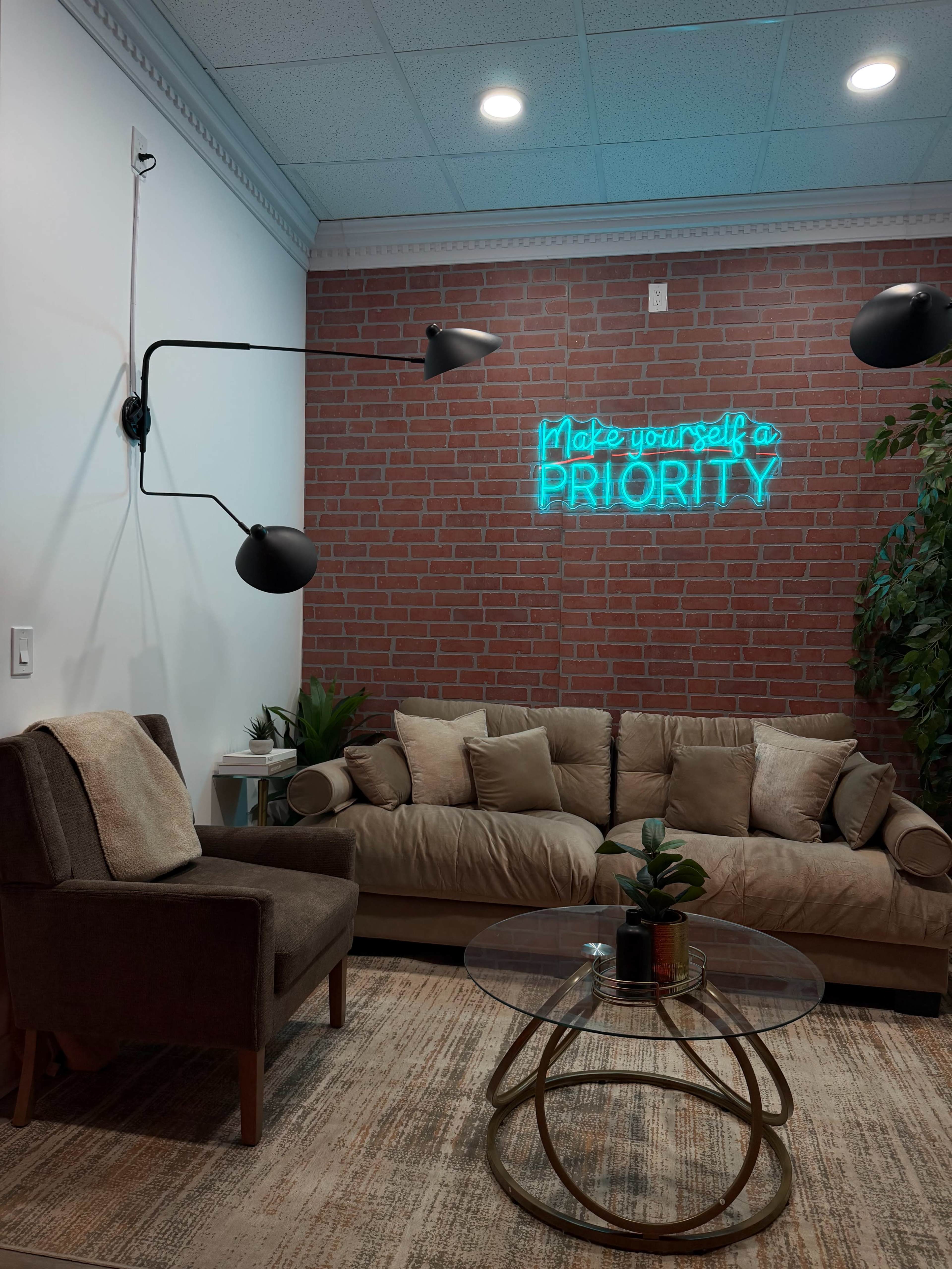 The image shows a cozy lounge area featuring a brown sofa, a glass coffee table with a small plant, a modern armchair, and a neon sign on a brick wall that reads "Make yourself a PRIORITY."