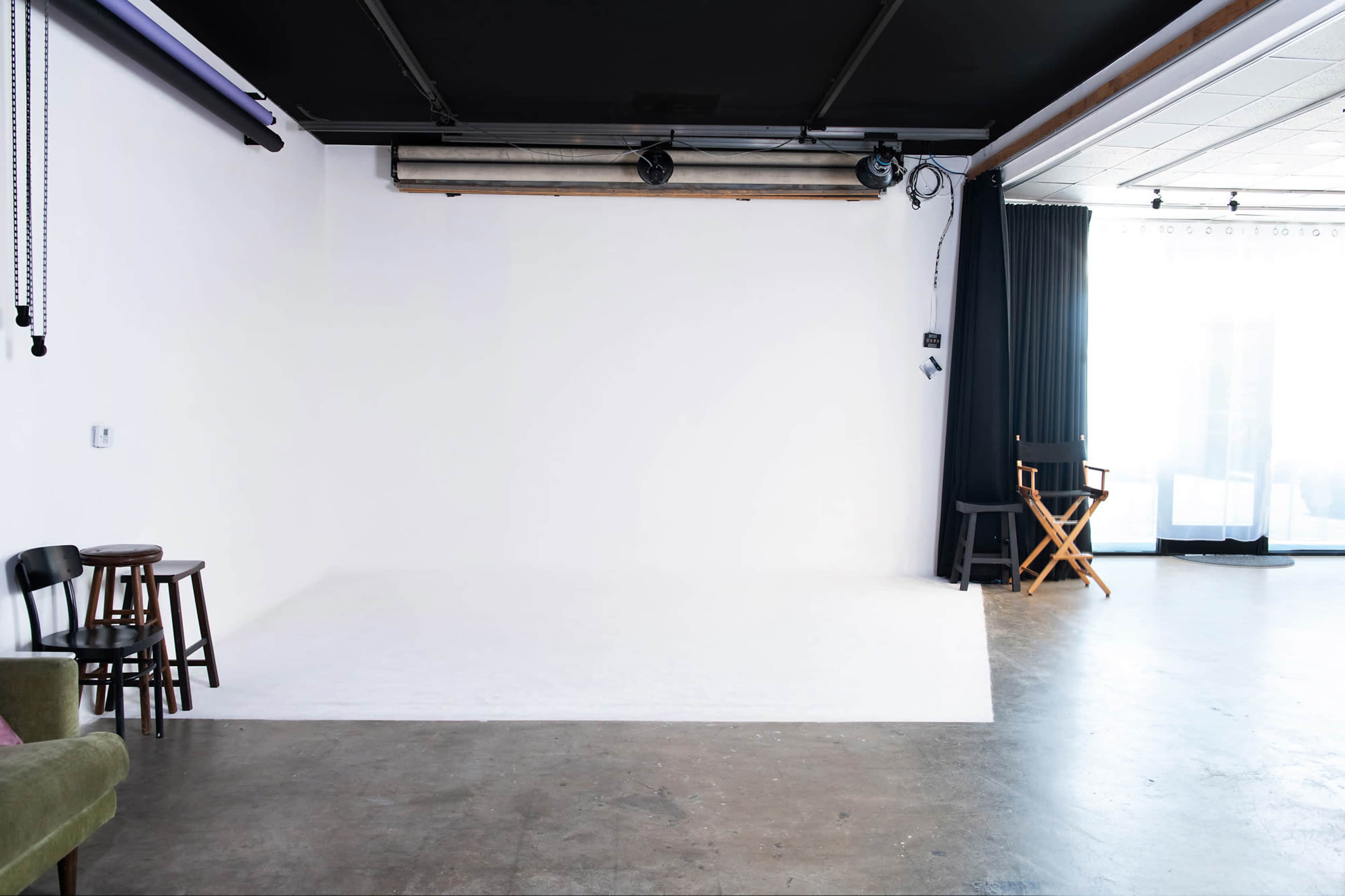 A minimalist photography studio with a white backdrop, two stools, a green couch, and a director's chair beside a large window.