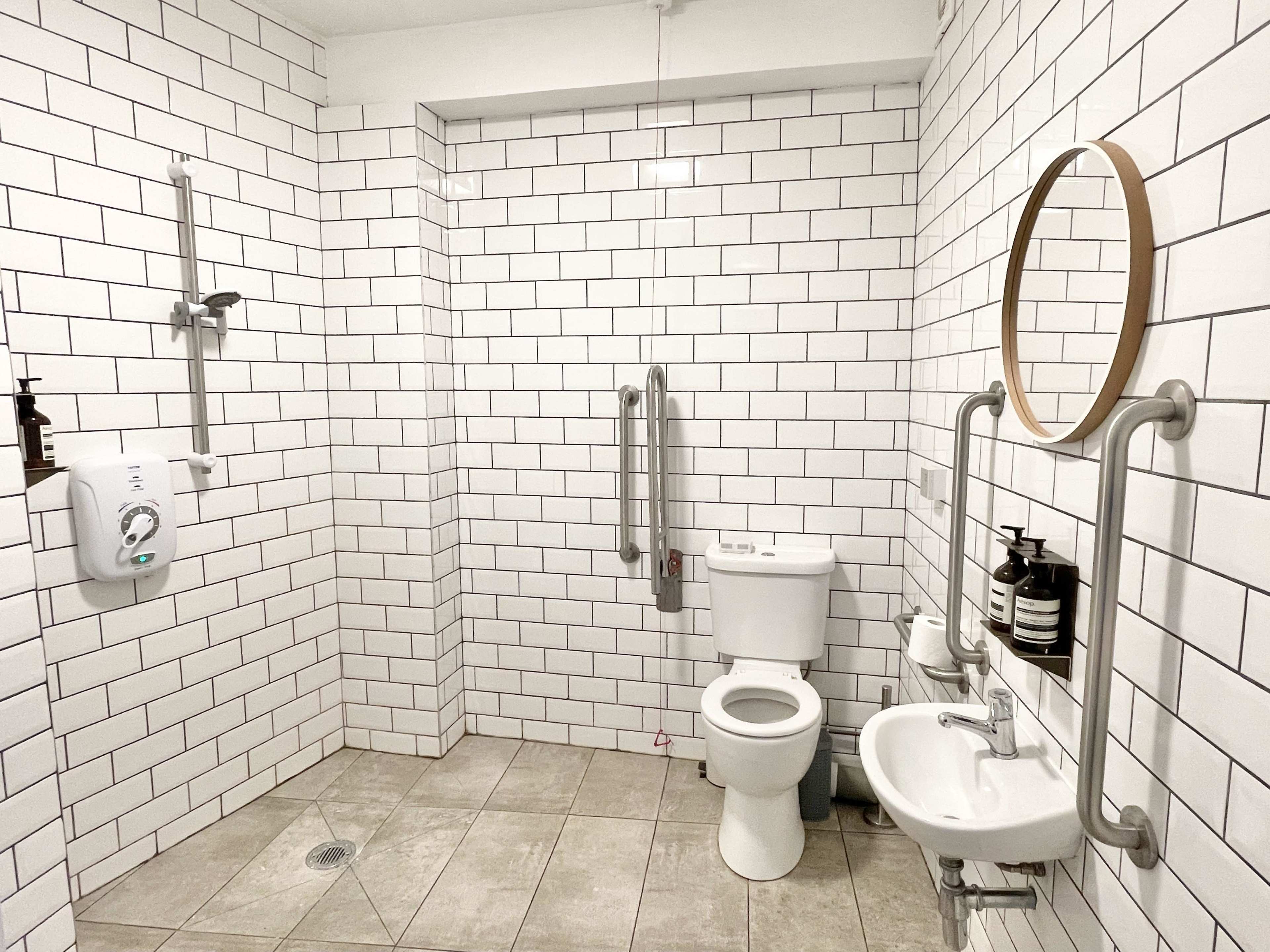 The image shows a modern bathroom with white tiled walls, featuring a toilet, sink, wall-mounted grab bars, and a shower with a handheld fixture.