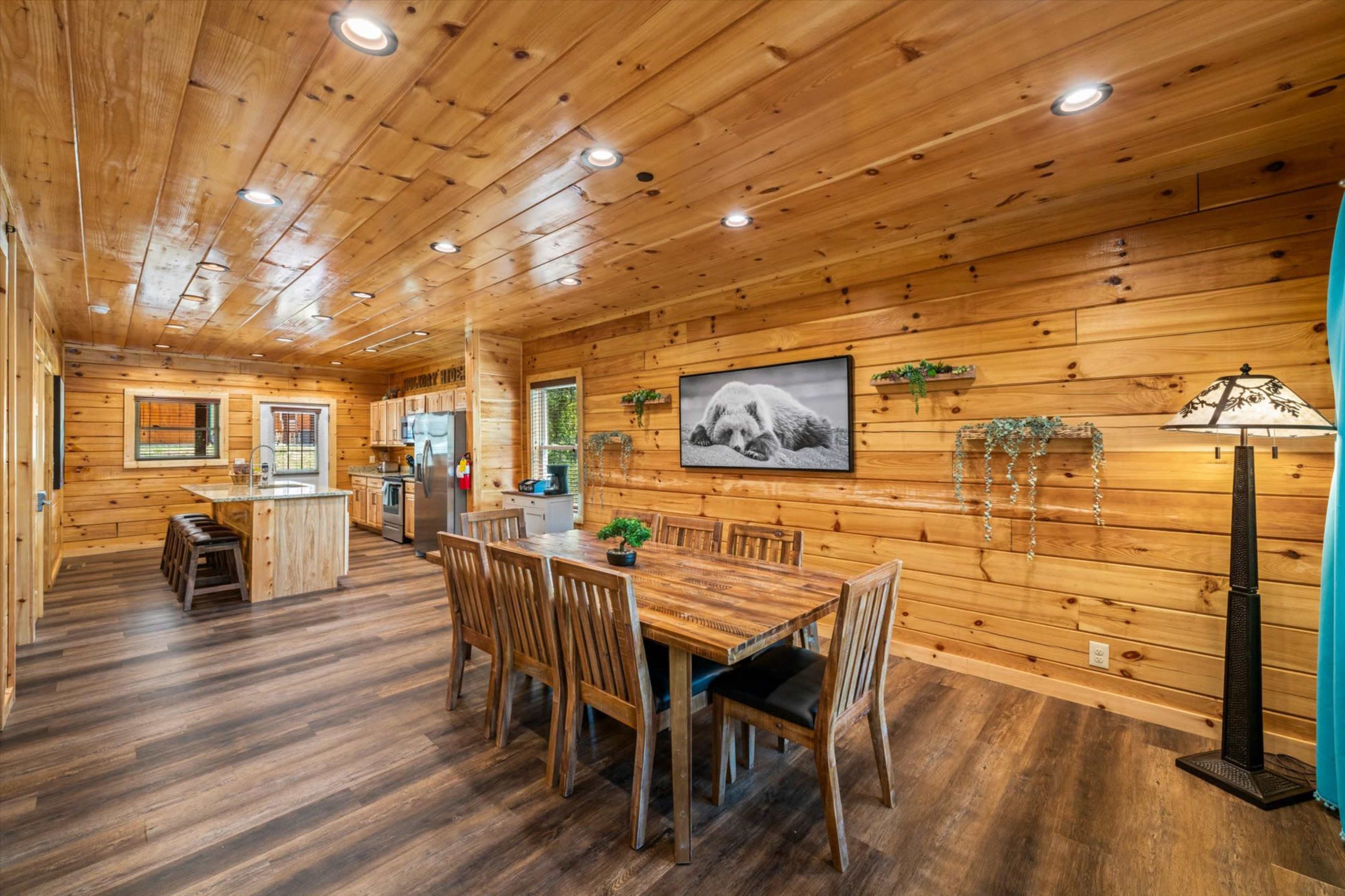A wooden interior of a cabin with a dining area, a kitchen, and a living space featuring a table and chairs, along with natural wood paneling on the walls and ceiling.