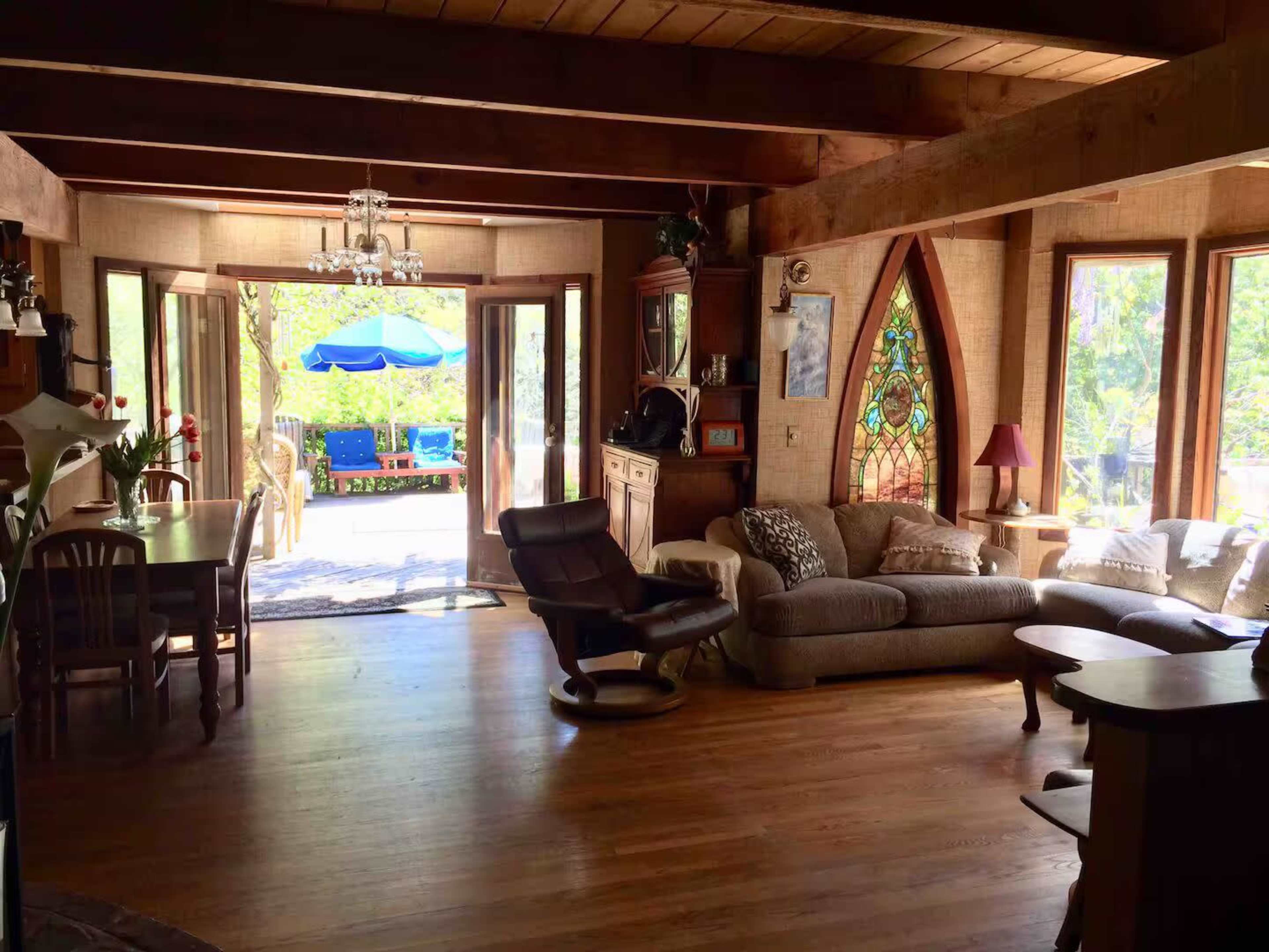 The image shows a cozy living room with wooden beams, a plush sofa, and large windows leading to a patio with outdoor seating and an umbrella.